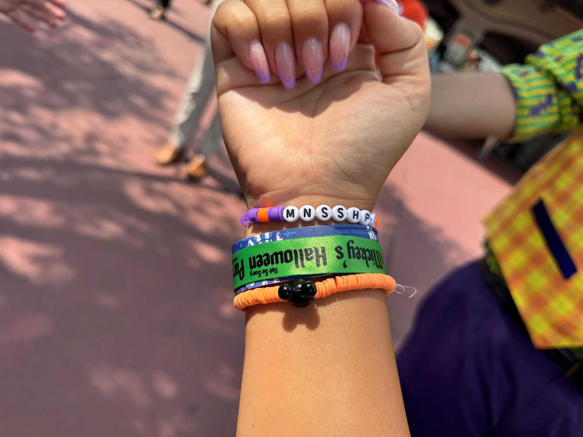A close-up of a person's wrist wearing several colorful bracelets, including one with beads spelling "MNSHPR," and a green bracelet reading "Mickey's Not-So-Scary Halloween Party.