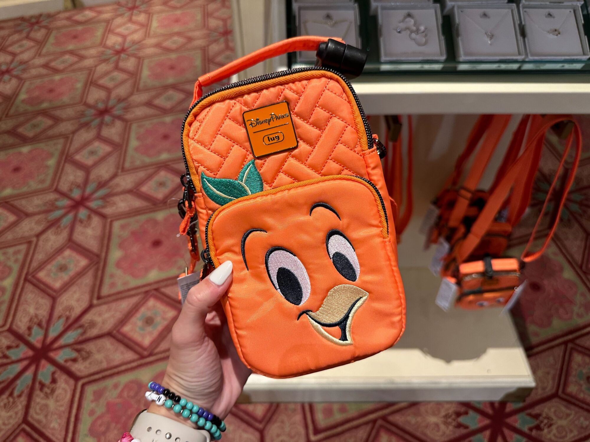 A hand holding an orange quilted bag with a cartoon orange face. Jewelry and identical bags are displayed in the background.