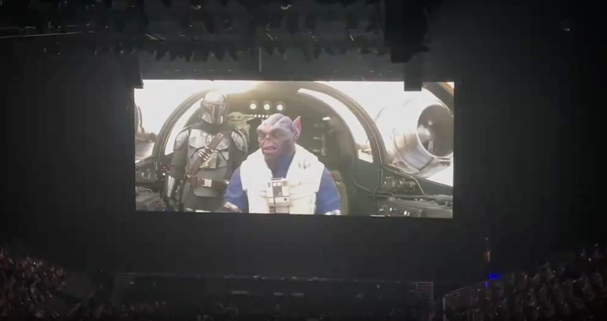 A large screen displays a scene from "The Mandalorian" featuring two characters in a spaceship cockpit. The scene is being watched by an audience in a darkened venue.