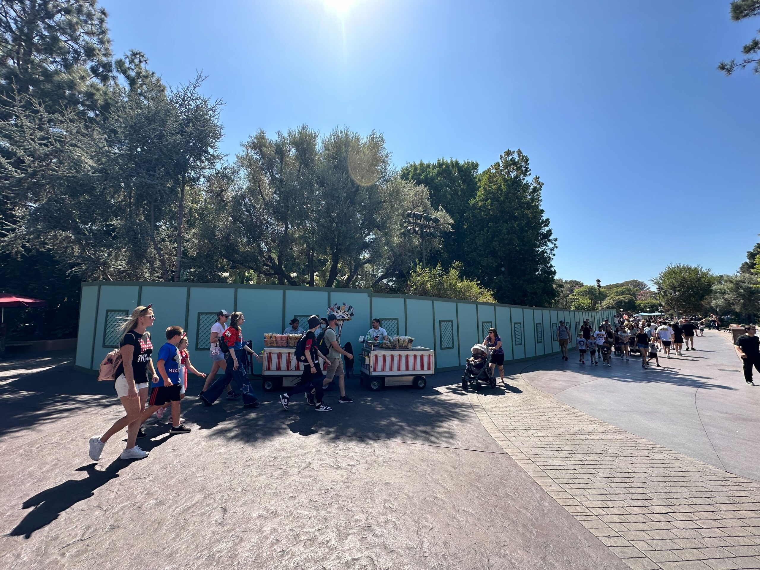 People walking past a construction site with green barriers and a food cart in a sunny outdoor park area.