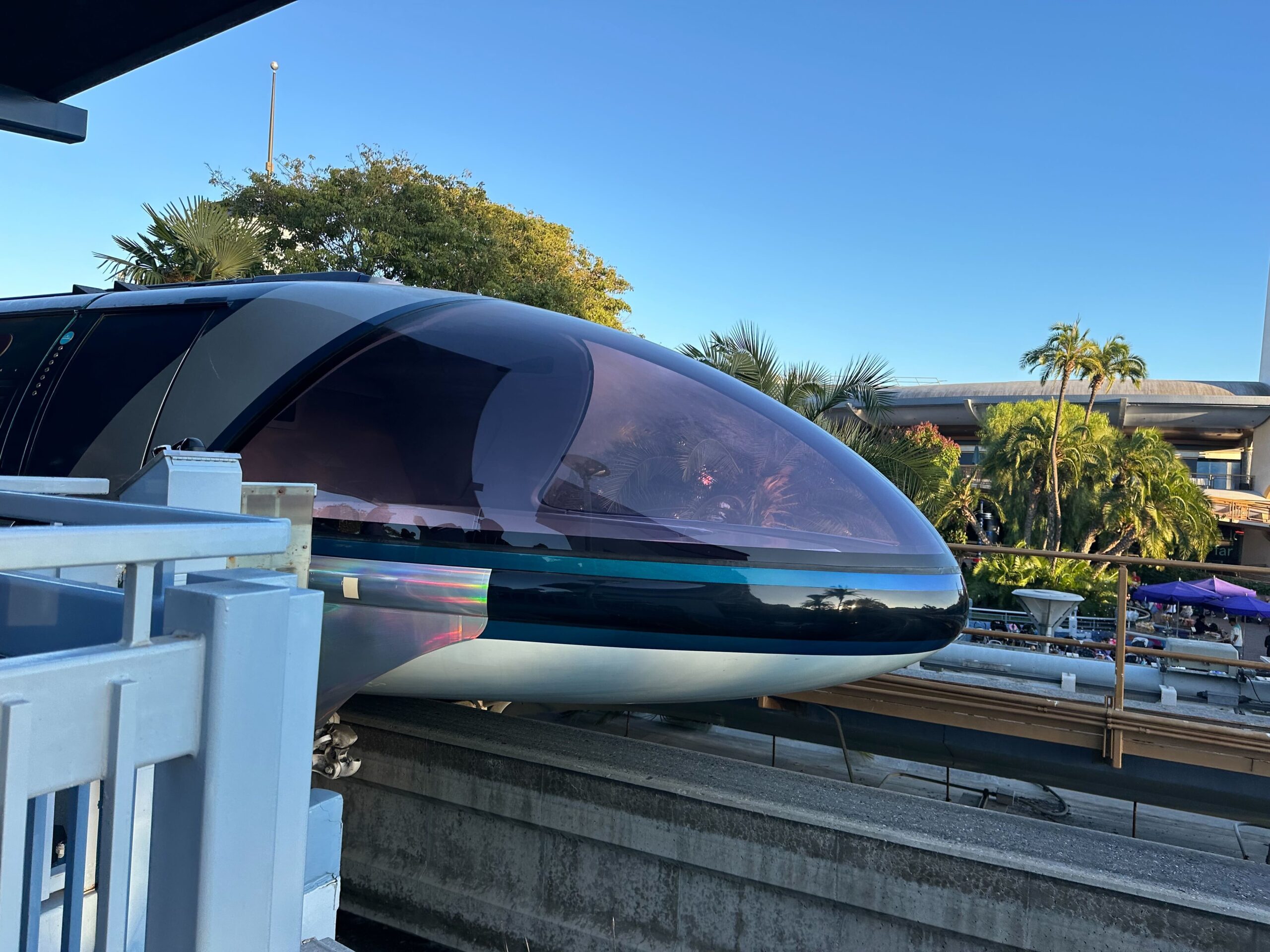 A futuristic Disneyland Monorail train with a sleek, aerodynamic front is at a station surrounded by palm trees and a clear blue sky, ready to welcome guests who might even choose to ride in the back for a unique experience.