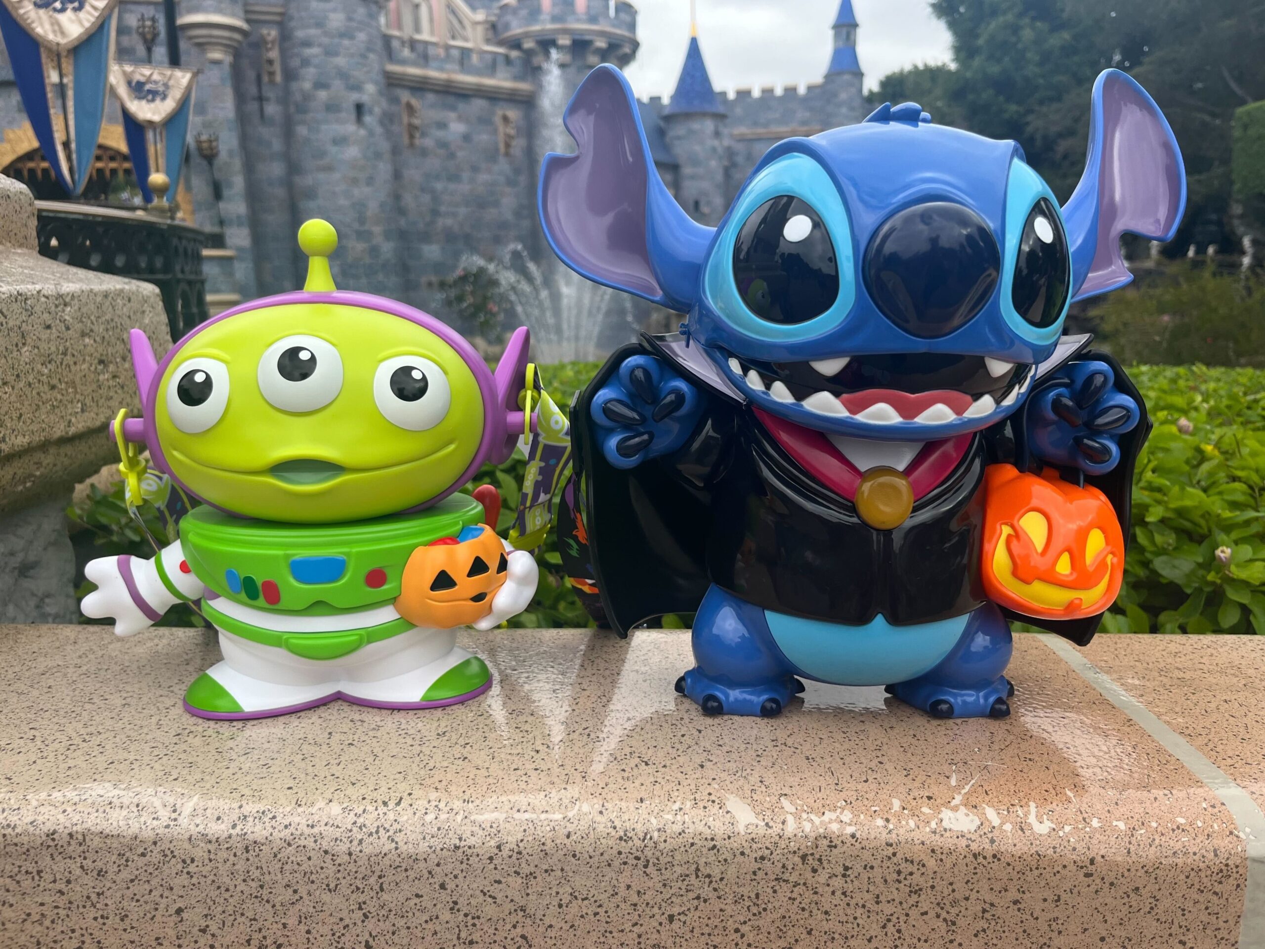 Two figures in costume: a three-eyed green alien holding a small pumpkin and a blue character dressed as a vampire with a jack-o'-lantern bucket, displayed in front of a castle backdrop.