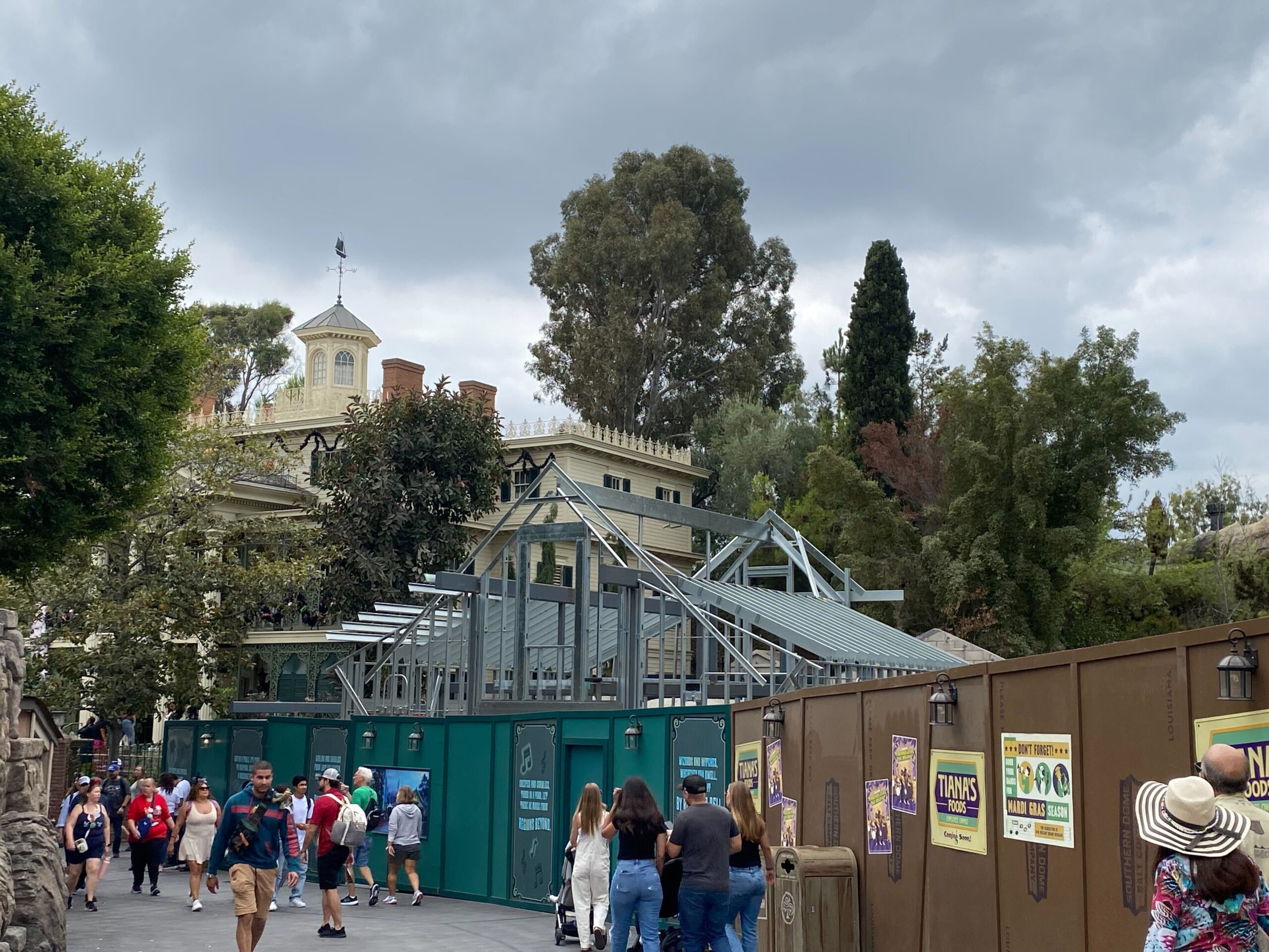 A group of people walk past a construction site surrounded by green and brown fences. The site has a partially built metal frame structure in front of a large building.