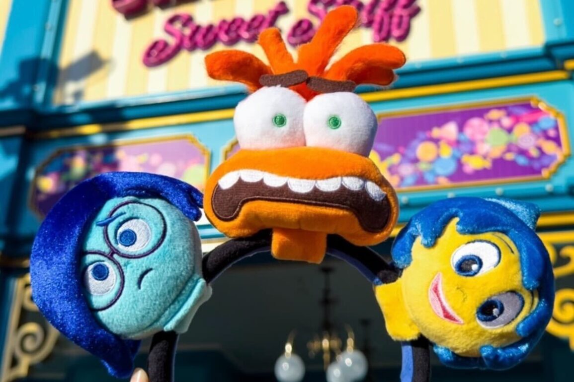 Three colorful cartoon character plush toys held up in front of a vibrantly decorated storefront. The characters include one with blue glasses looking sad, one with wide eyes and orange hair, and one smiling.