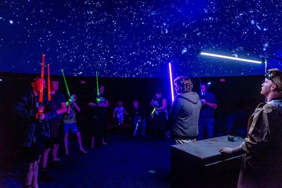 People participating in a lightsaber training event under a dome with a starry sky projection.
