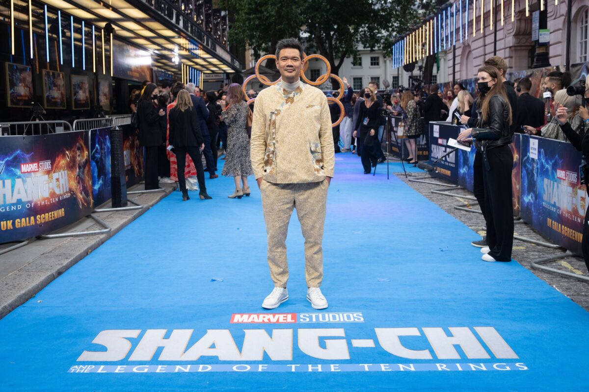 A person stands on the blue carpet at the UK Gala Screening of "Shang-Chi and the Legend of the Ten Rings," directed by Destin Daniel Cretton, wearing a beige outfit and white sneakers, surrounded by attendees and photographers.