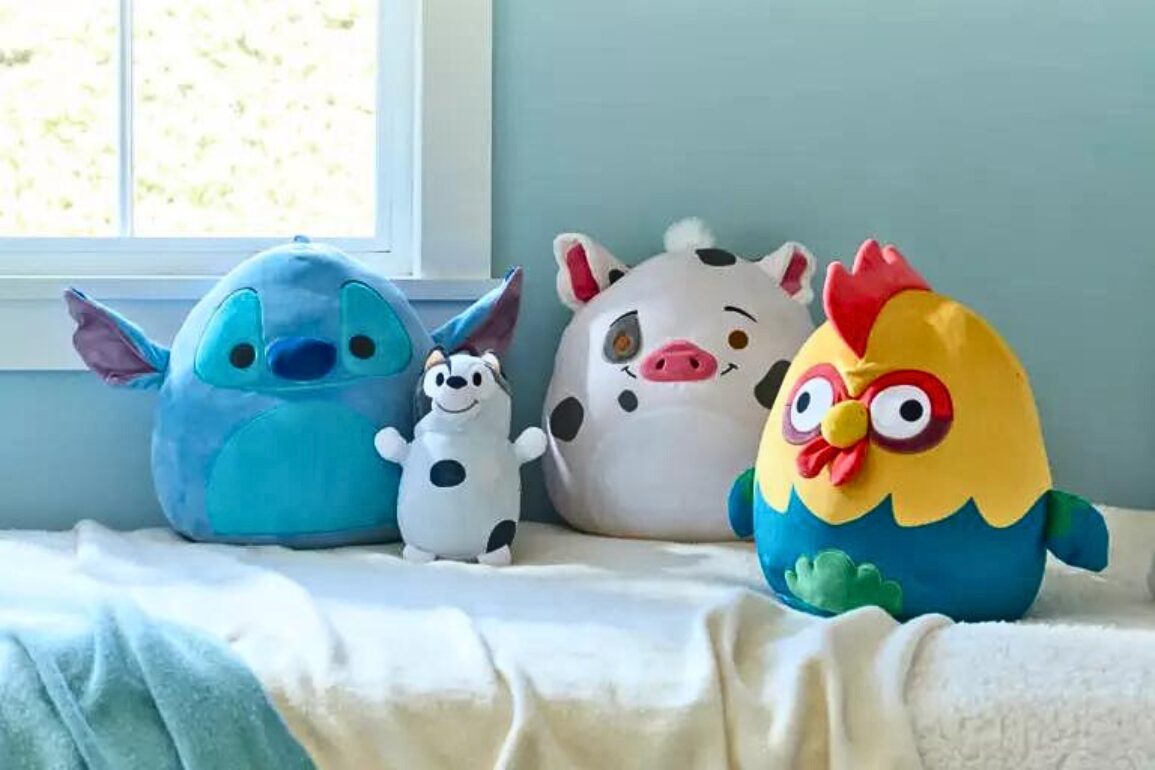 Four squishmallows, including blue, white, pig, and chicken characters, are placed on a light-colored blanket near a window.