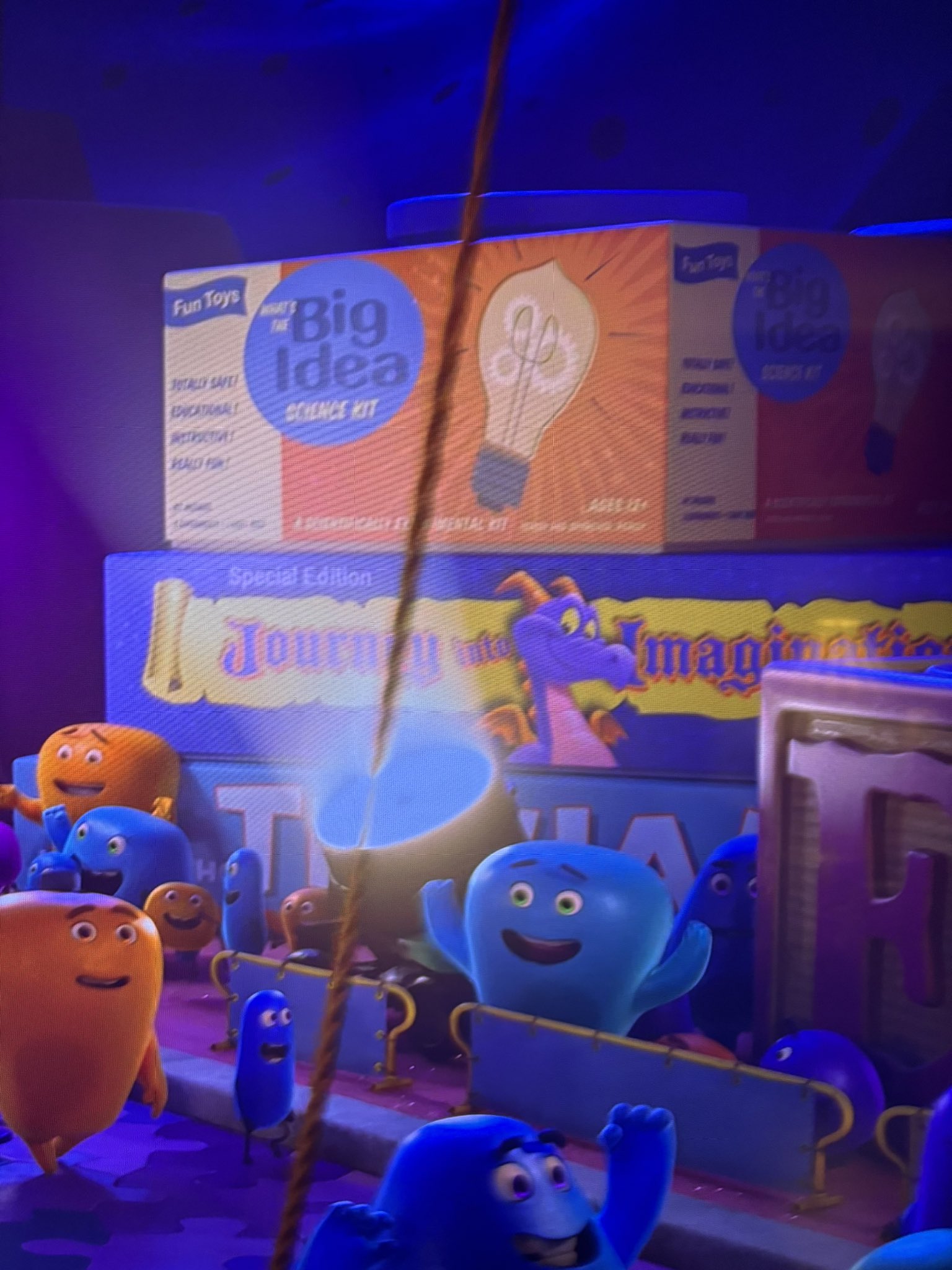 Colorful animated characters gather around various toys and kits, including a "Big Idea Science Kit" and a "Journey into Imagination" book featuring Figment, all set against a playful, vibrant backdrop with hidden Easter eggs from Inside Out 2.