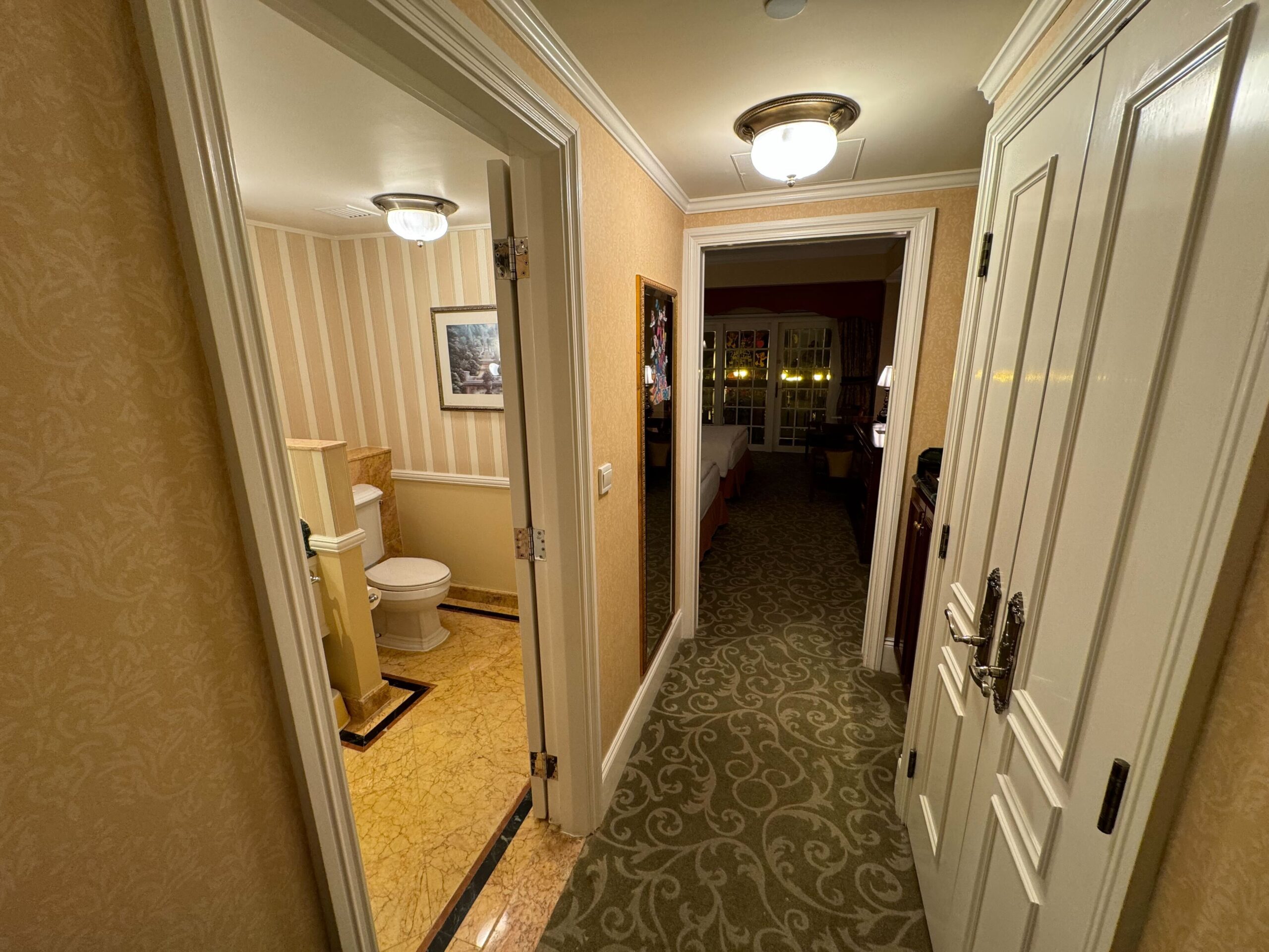 A hallway in the Hong Kong Disneyland Hotel showcases a bathroom door on the left, featuring a toilet and framed artwork. An open door leads to a Deluxe Room with carpeted flooring, elegant furniture, and warm lighting. The highlight is the balcony offering stunning views.