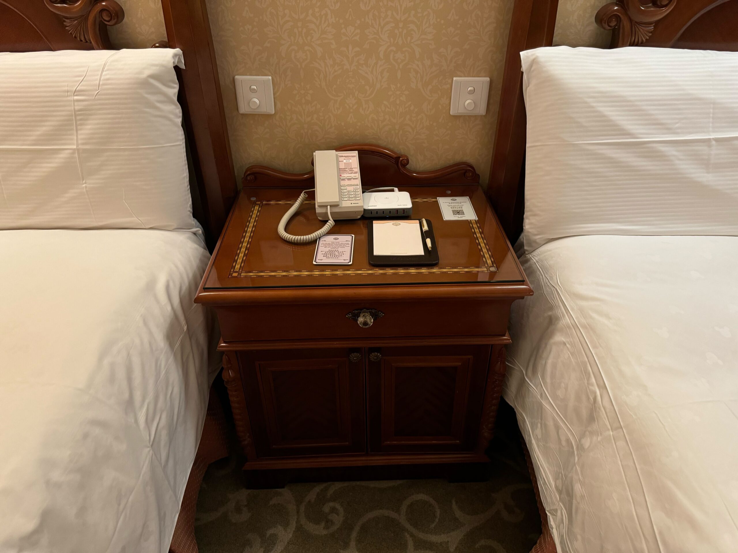 A wooden nightstand with a phone, clock, and notepads on top is positioned between two neatly made beds with white linens in the Deluxe Room at Hong Kong Disneyland Hotel. The room also features a charming balcony overlooking the magical surroundings.