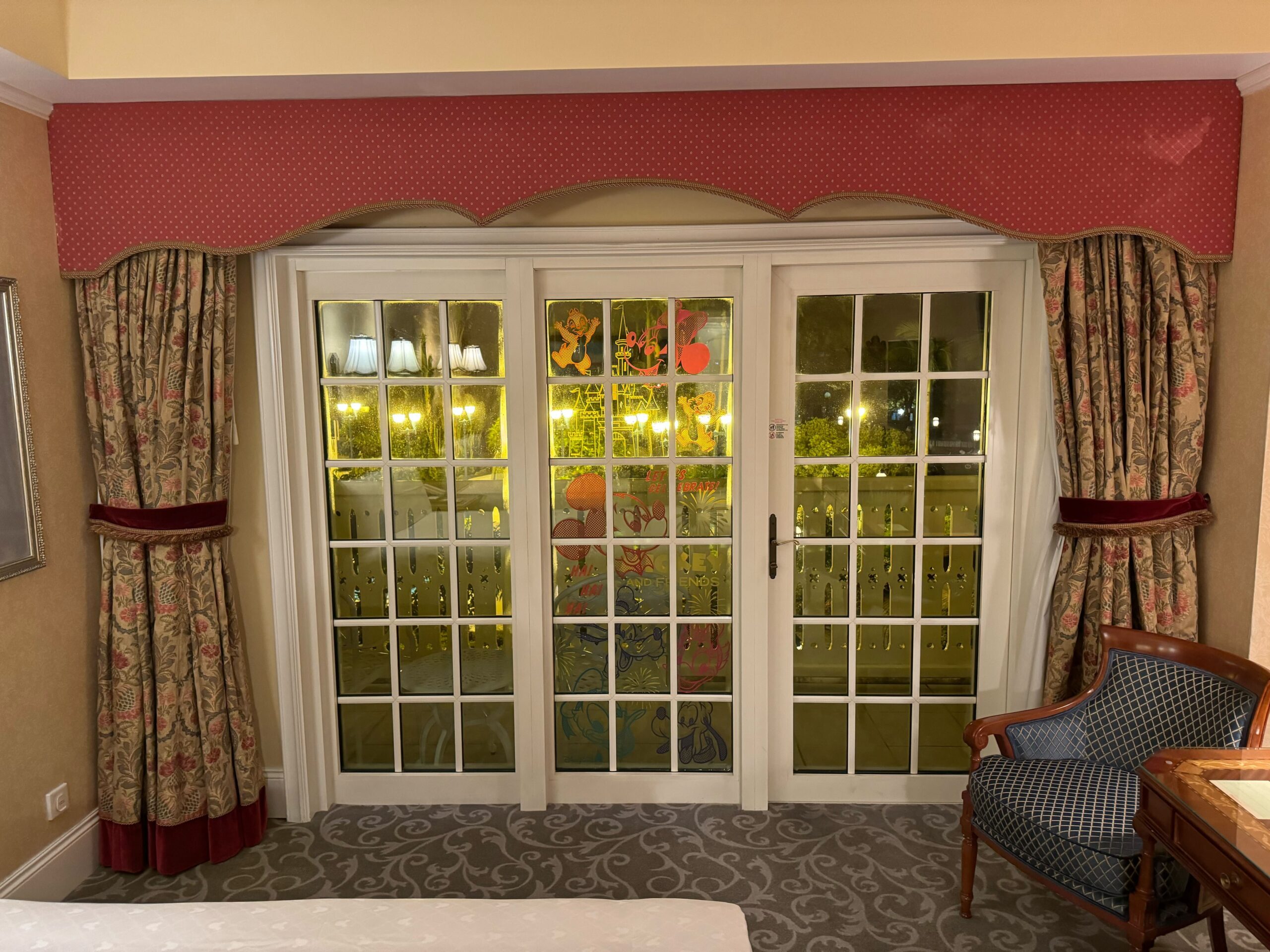 A brightly lit Deluxe Room at the Hong Kong Disneyland Hotel features French doors with patterned curtains, a chair to the right, and a view of the balcony or garden through the glass panels.
