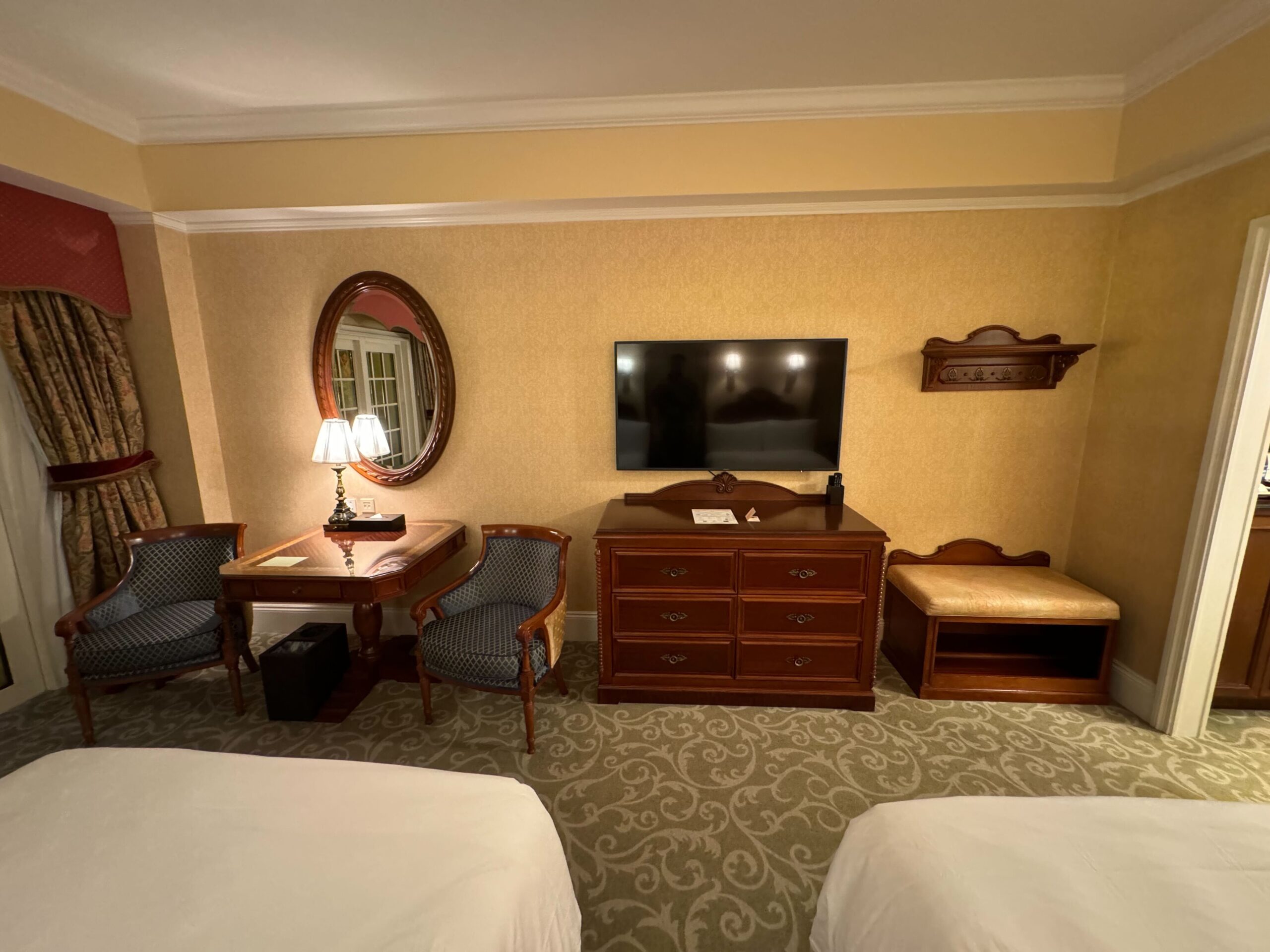 A Deluxe Room at the Hong Kong Disneyland Hotel features a dresser with a mounted flat-screen TV, a small dining set with two chairs, a wall mirror, and a cushioned bench. A partially visible bed is in the foreground, while the balcony offers enchanting views of the surroundings.
