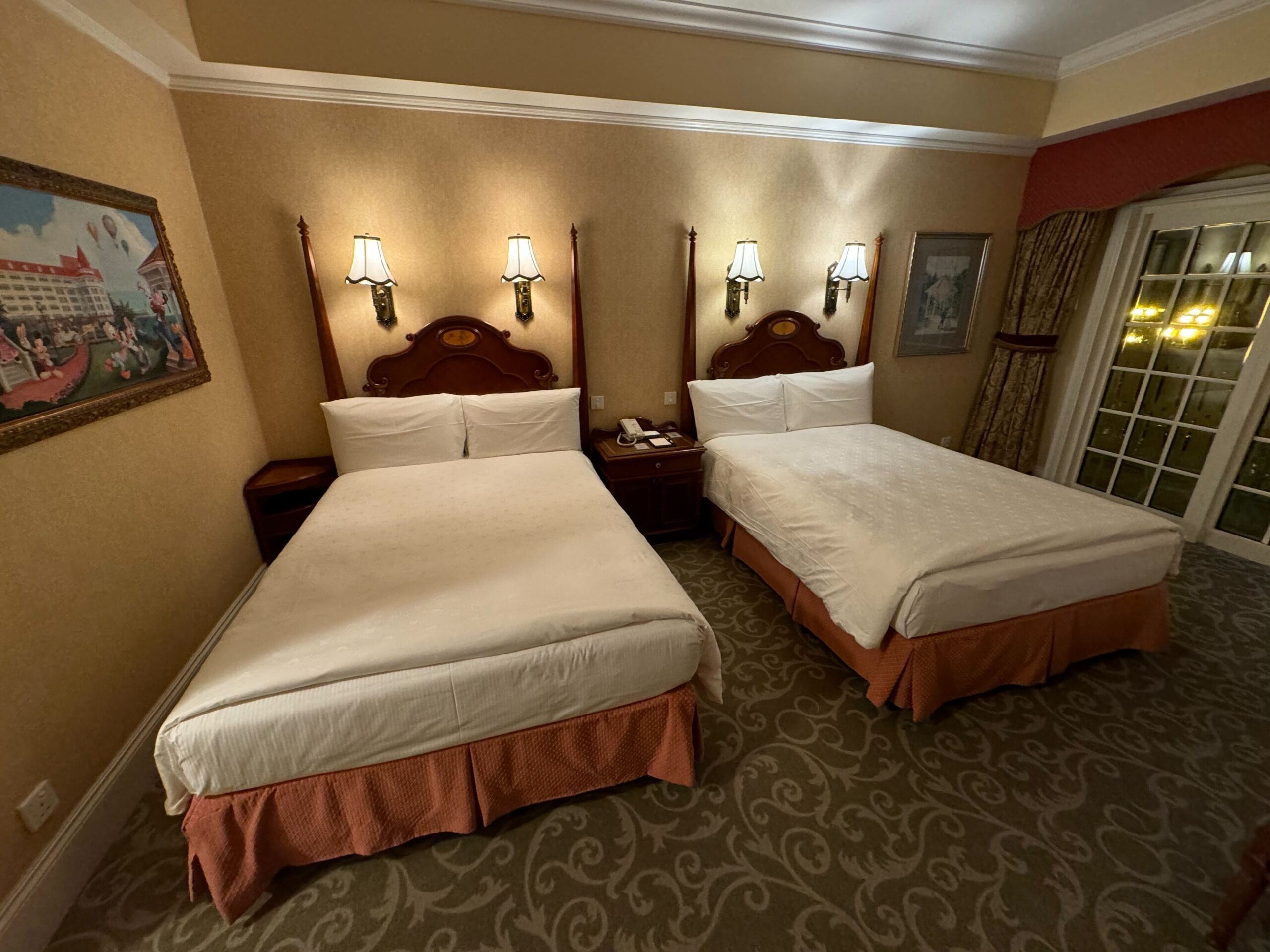 A deluxe room at the Hong Kong Disneyland Hotel features two double beds with white linens, wall-mounted lamps, wooden headboards, decorative wall art, nightstands, and a glass door leading to a balcony or another room.