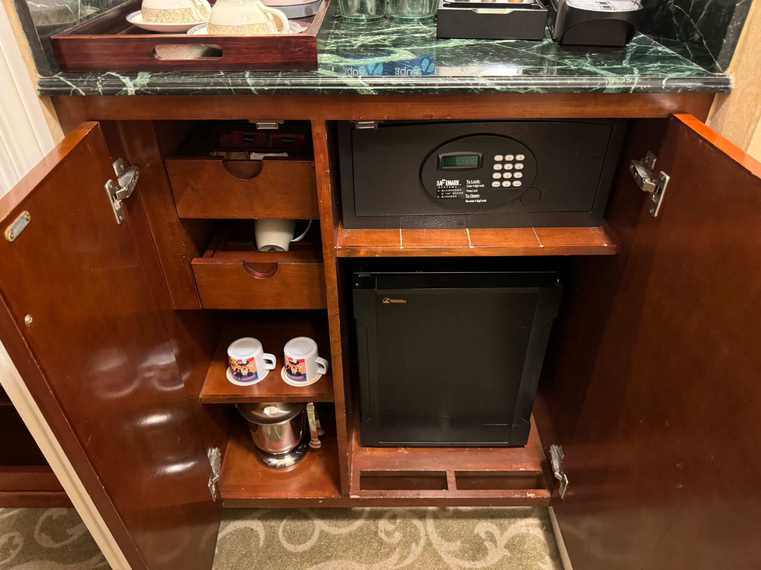 Discover the charm of our Deluxe Room at Hong Kong Disneyland Hotel. The hotel room cabinet, with open doors, reveals a small refrigerator, a safe, two coffee mugs with a coffee maker, and a few drawers. Enjoy the convenience and luxury during your stay.