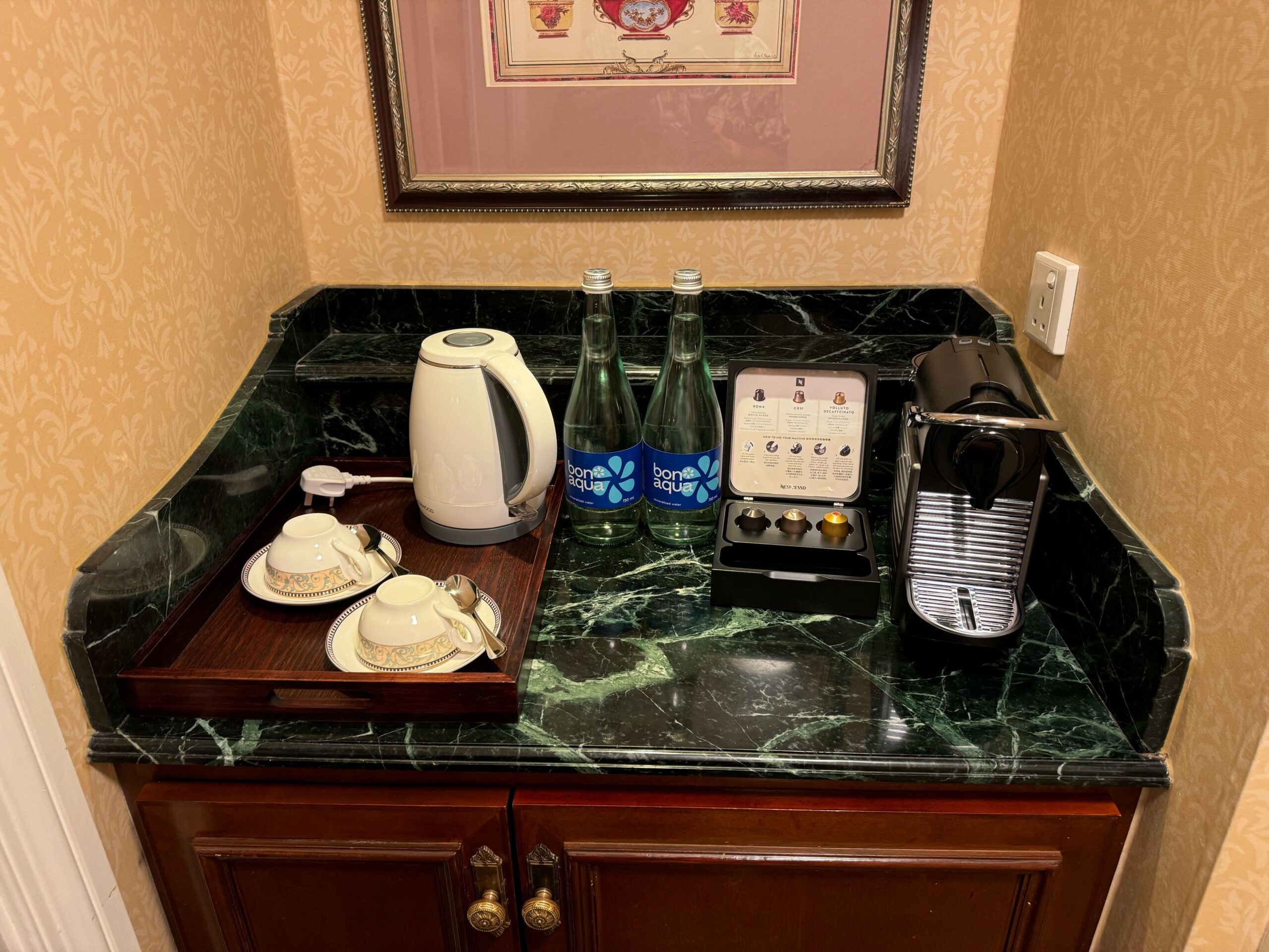 A small countertop in the Deluxe Room at Hong Kong Disneyland Hotel features a coffee setup with a Nespresso machine, a kettle, two cups and saucers on a tray, and two bottles of water. Enjoy your morning brew before stepping out onto the balcony for a breath of fresh air.