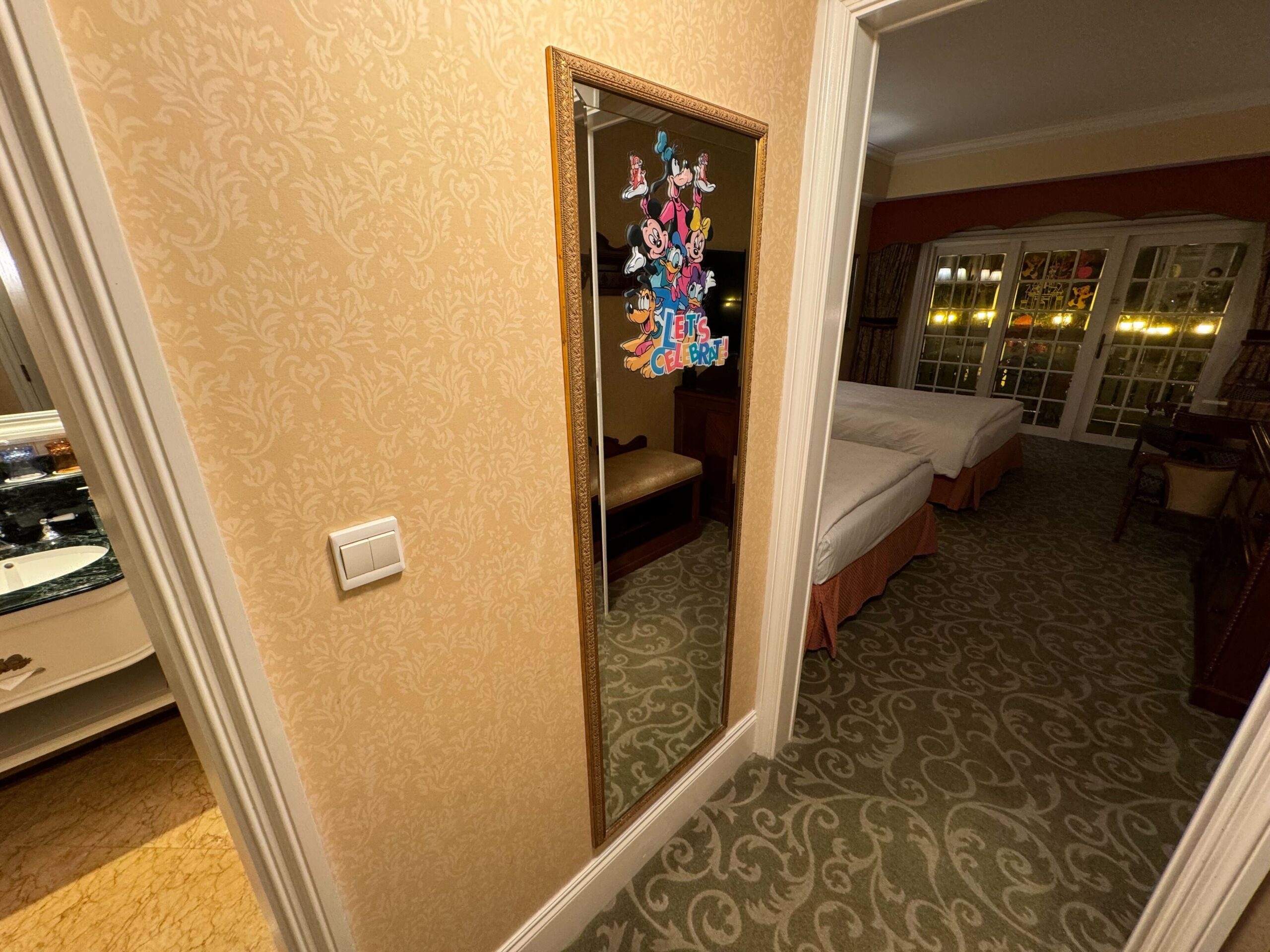 A hotel room hallway with a full-length mirror decorated with colorful stickers and a "Let's Celebrate" sign. The Deluxe Room has two beds, glass doors leading to another area, and a balcony with views reminiscent of the Hong Kong Disneyland Hotel.