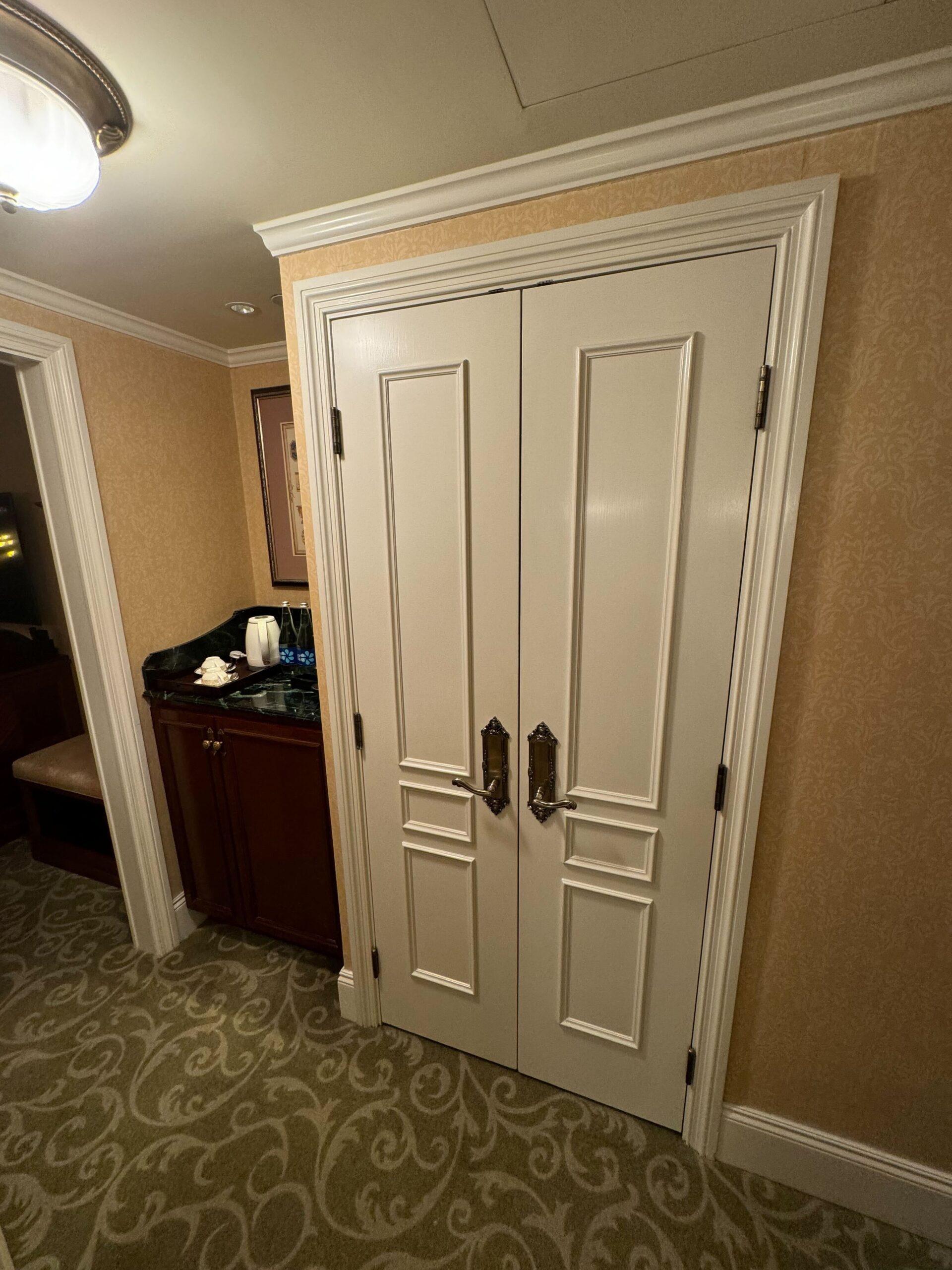 A double white door with ornate handles stands in a room with patterned carpet and beige walls. To the left, a small dark wood cabinet holds various items, and a framed picture hangs above it, capturing the elegance reminiscent of a Deluxe Room at the Hong Kong Disneyland Hotel.