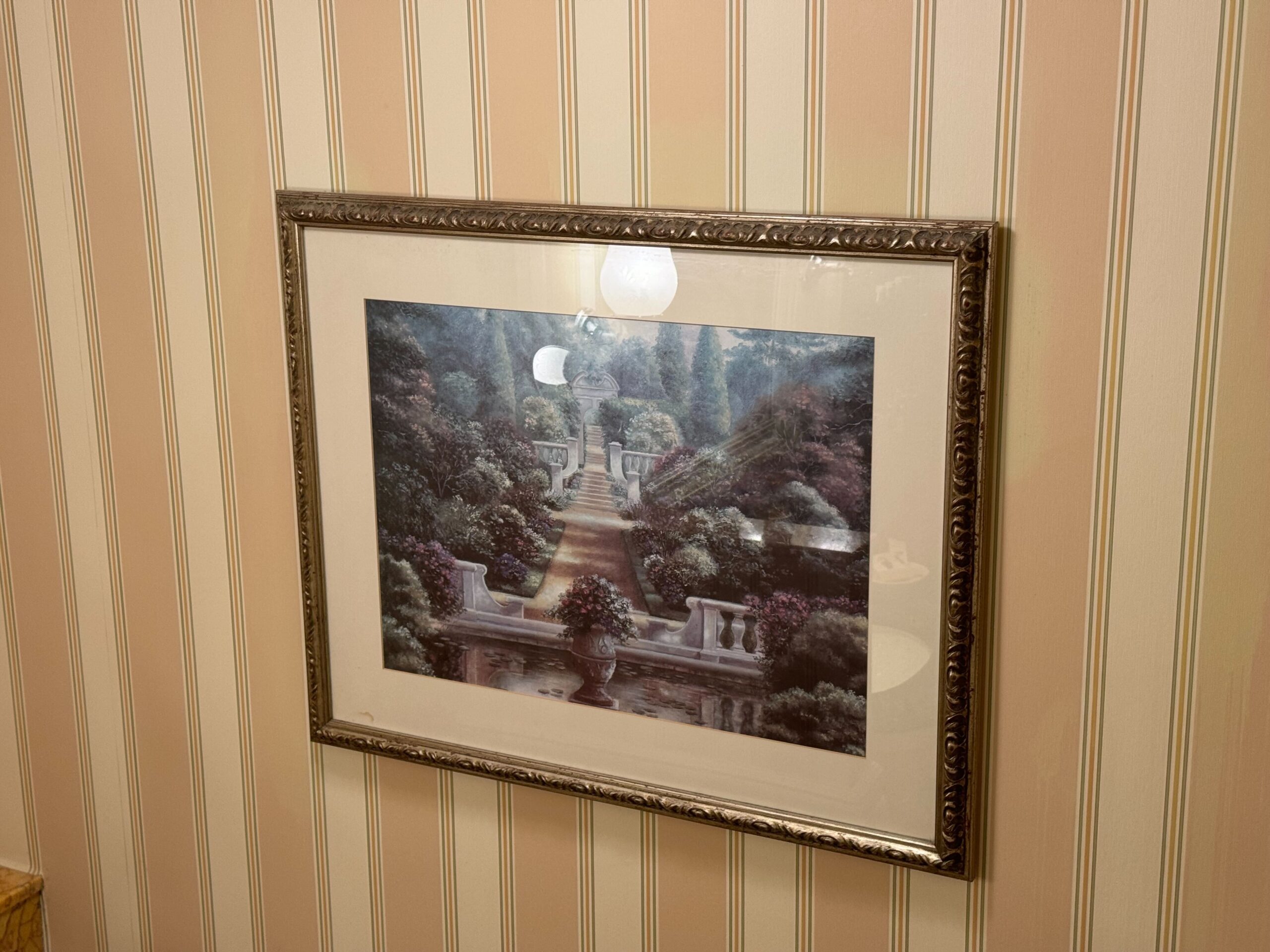 A framed painting of a garden pathway with trees and plants hangs on a wall with beige striped wallpaper, adding charm to the Deluxe Room at the Hong Kong Disneyland Hotel.