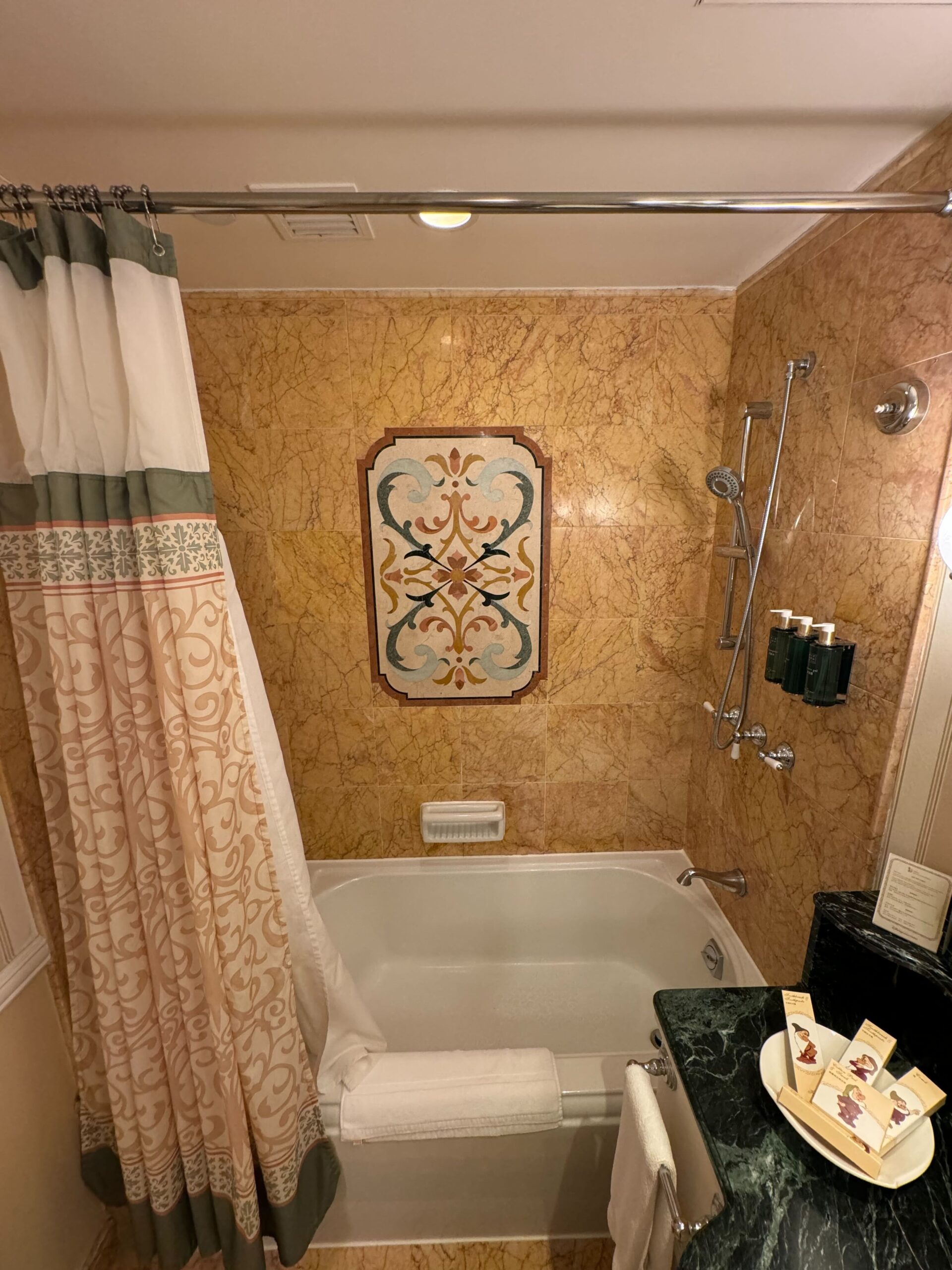 Image of a small bathroom in a Deluxe Room at the Hong Kong Disneyland Hotel, featuring a bathtub with a floral-patterned shower curtain and decorative tile on the wall. A glass shelf holding toiletries is mounted above the bathtub, and a towel is draped over the tub.