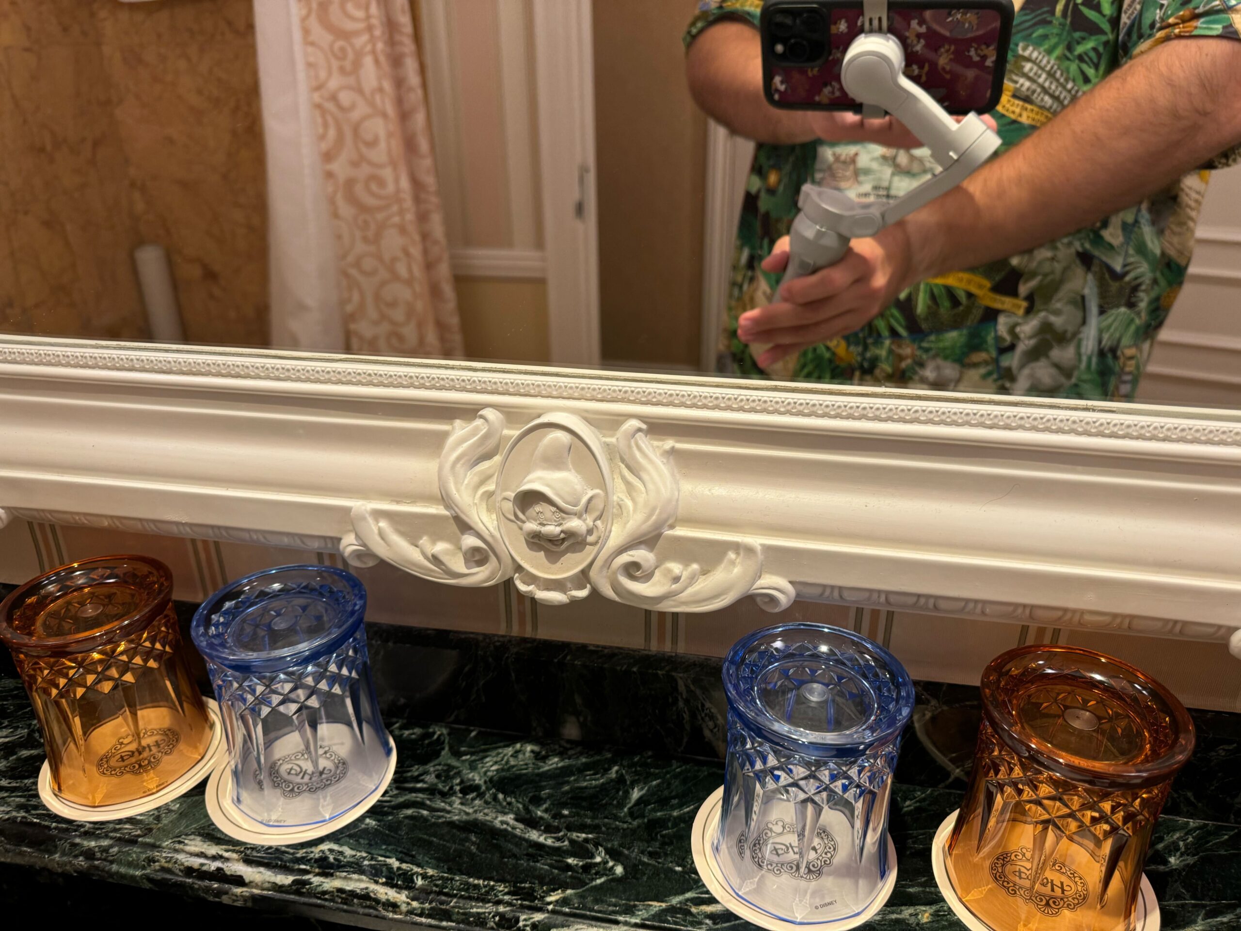 A person taking a selfie in a mirror, wearing a green shirt with a floral pattern, stands in what looks like the Deluxe Room of the Hong Kong Disneyland Hotel. Three decorative drinking glasses in yellow, blue, and brown are placed on a marble counter.