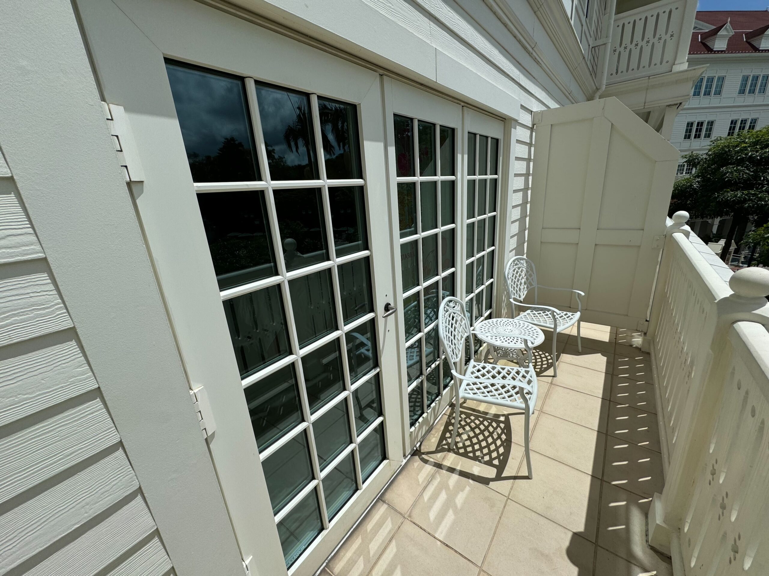 A sunlit balcony with white French doors, two white metal chairs, and a matching table. This quaint space is part of a Deluxe Room at the Hong Kong Disneyland Hotel and overlooks a courtyard with other white buildings visible on the right.
