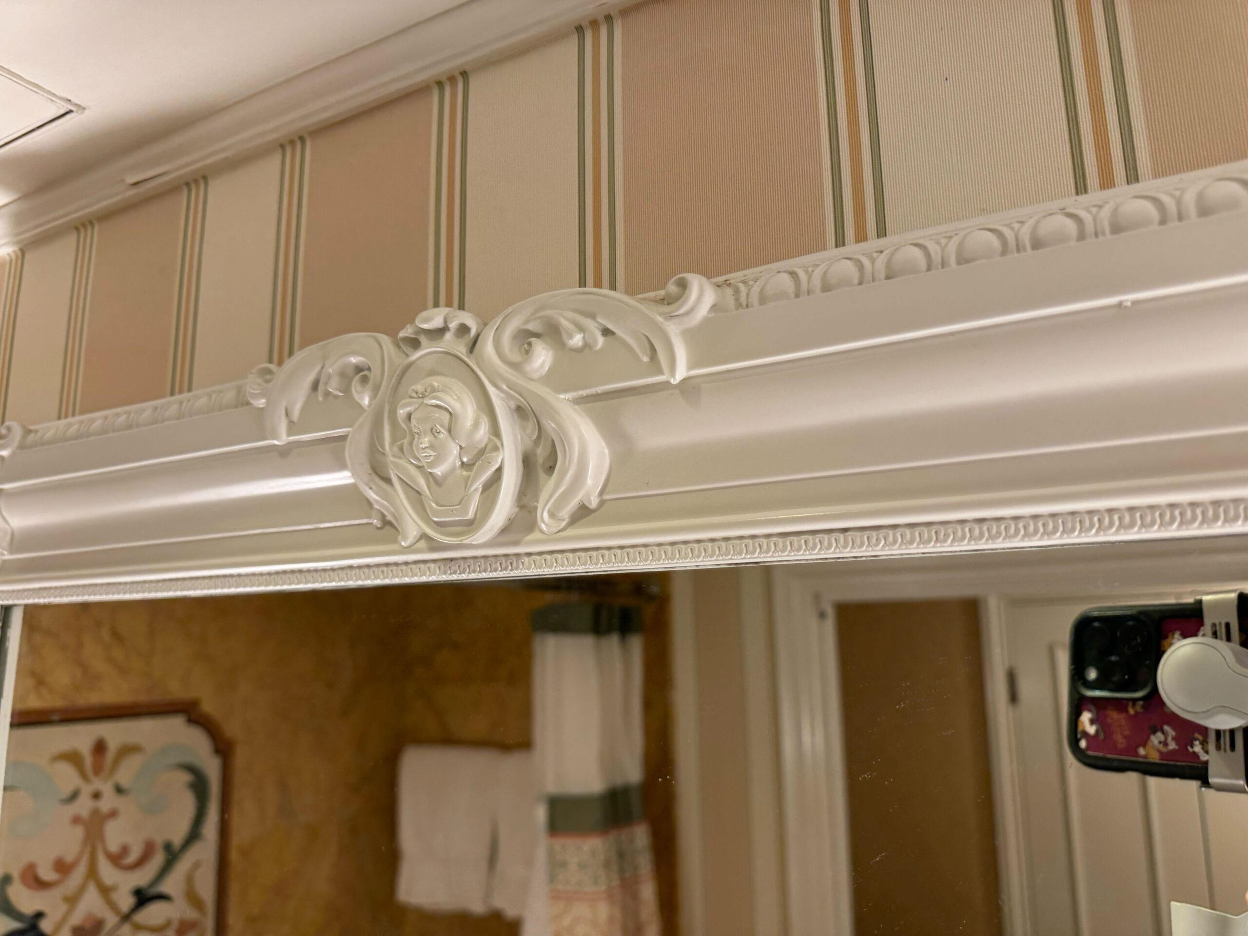 Top portion of a decorative bathroom mirror with a detailed white frame and a striped wallpaper background in a deluxe room. Part of the curtain, a towel, and a decorative wall art piece are also visible.