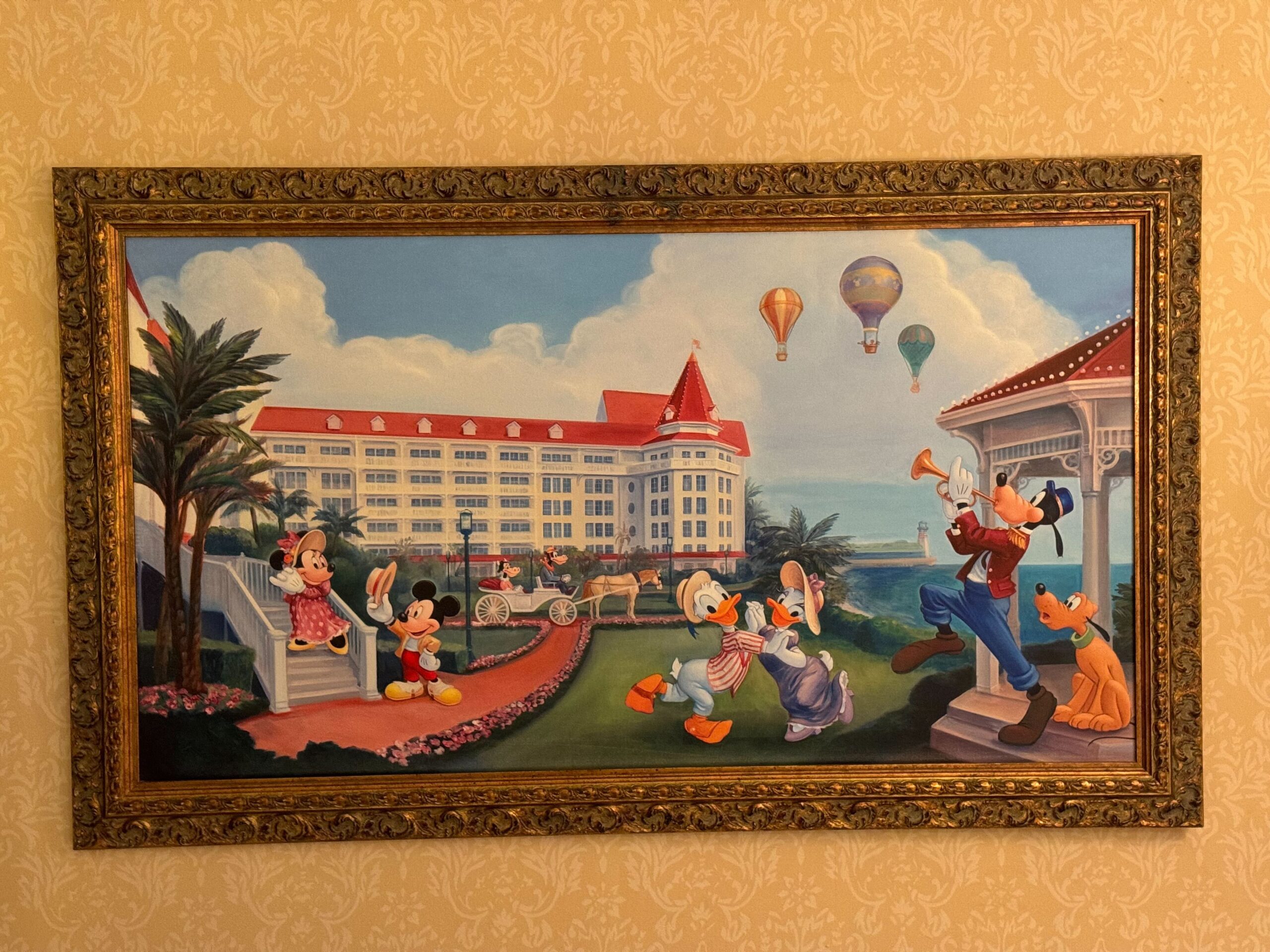 A framed painting depicts animated characters in a lively outdoor scene with the grand Hong Kong Disneyland Hotel, hot air balloons, and trees in the background, evoking the charm of a Deluxe Room's balcony view.
