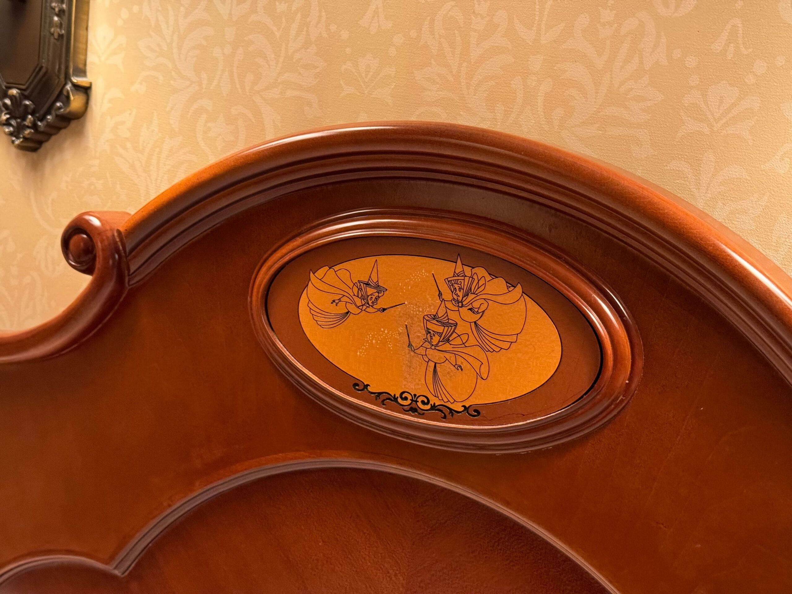 A wooden bed frame with an oval inset featuring an illustration of three animated fairy characters in the center, perfect for adding a touch of magic to your Deluxe Room at Hong Kong Disneyland Hotel.