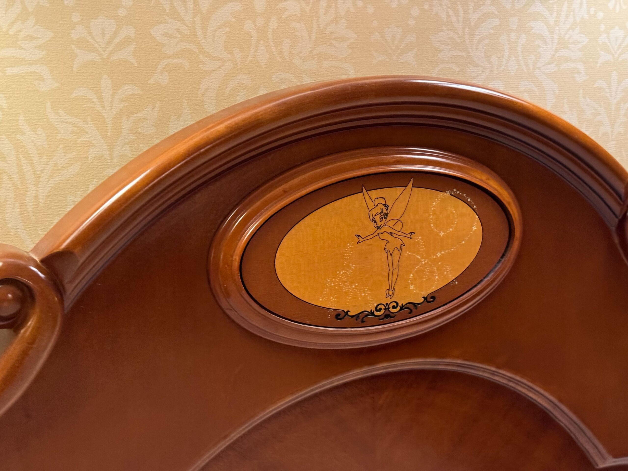 A wooden headboard with an oval inset featuring an image of a fairy with wings on a light brown background graces the Deluxe Room at Hong Kong Disneyland Hotel. The background wallpaper shows a subtle, decorative pattern, and the room opens to a charming balcony.