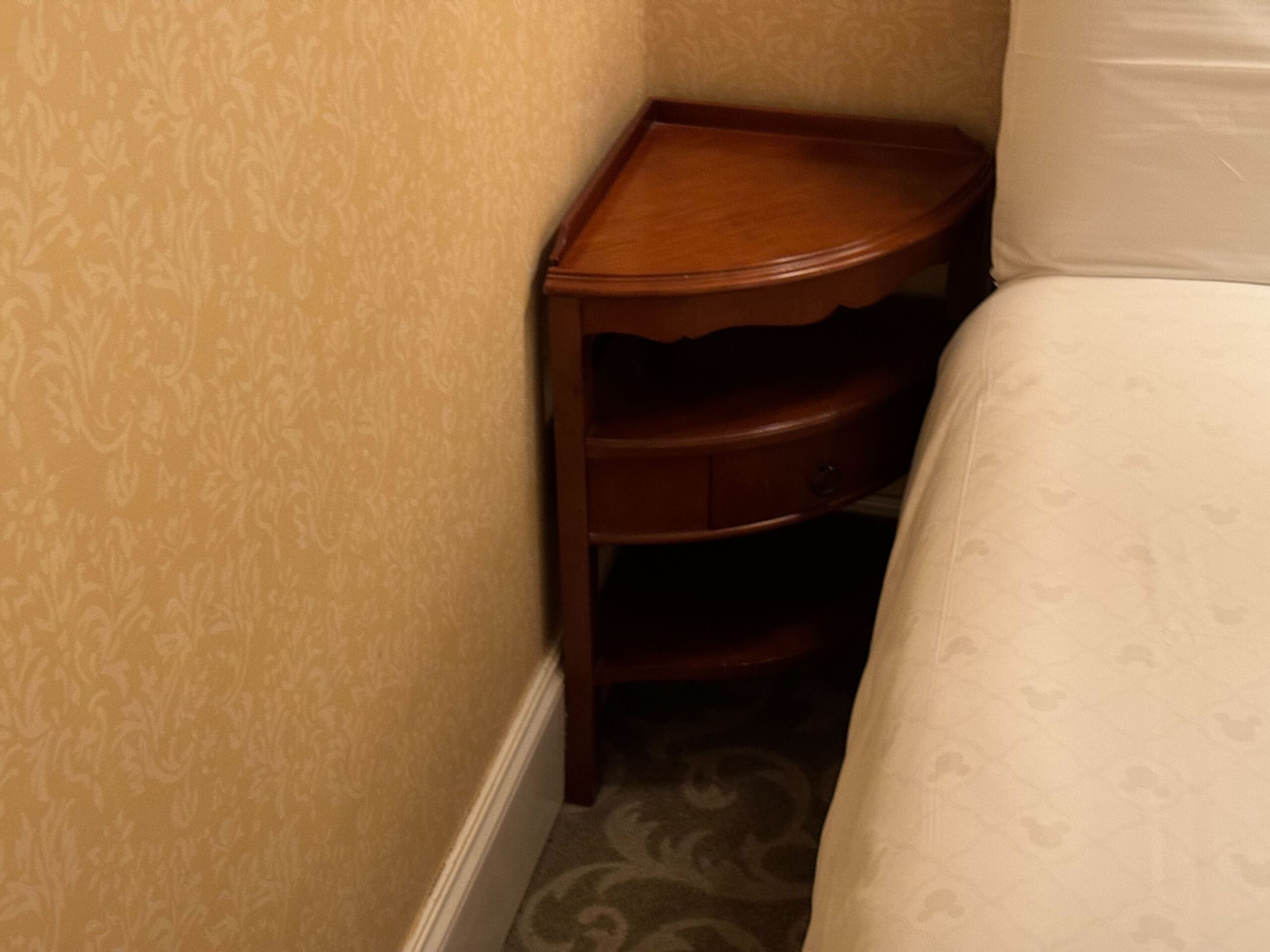 A small, wooden, triangular bookshelf stands between a patterned wallpapered wall and a bed with a white cover in the Deluxe Room of the Hong Kong Disneyland Hotel.
