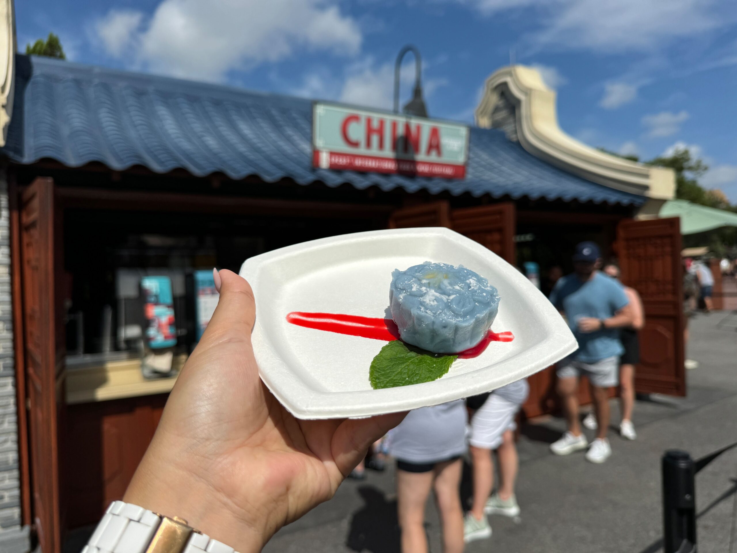 A hand holding a white plate with blue jelly dessert, reminiscent of a Snowy Mooncake, garnished with a mint leaf and red sauce in front of the China Booth at the EPCOT Food & Wine Festival.