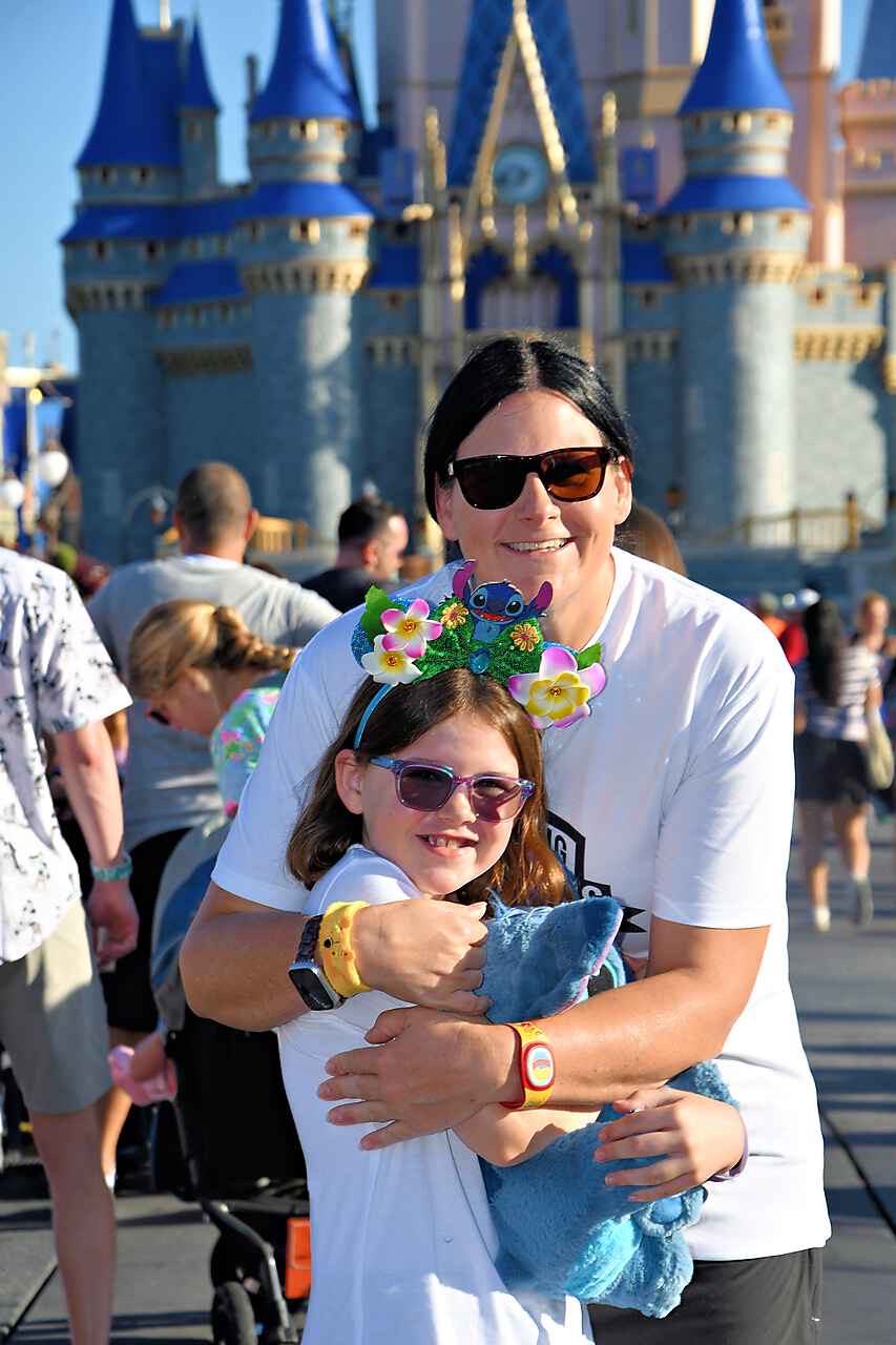 An adult and a child, both wearing sunglasses, stand hugging in front of a castle with blue spires. The child wears colorful floral headbands and holds a blue plush toy. Other visitors are in the background.