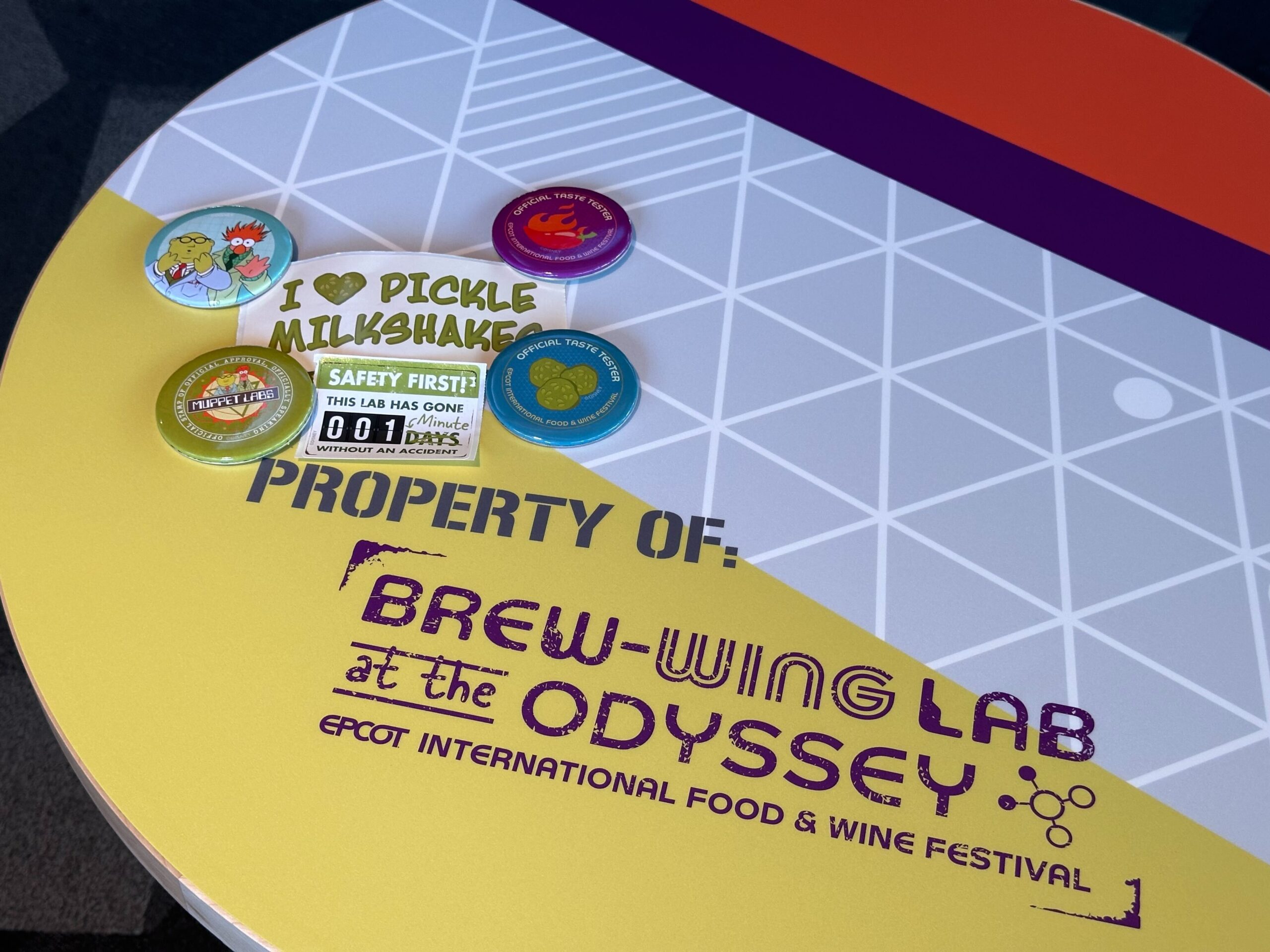 A table with "Brew-Wing Lab at the Odyssey" branding displays various colorful buttons, including one saying "I ♥ PICKLE MILKSHAKES," and a safety sign indicating zero lost days.