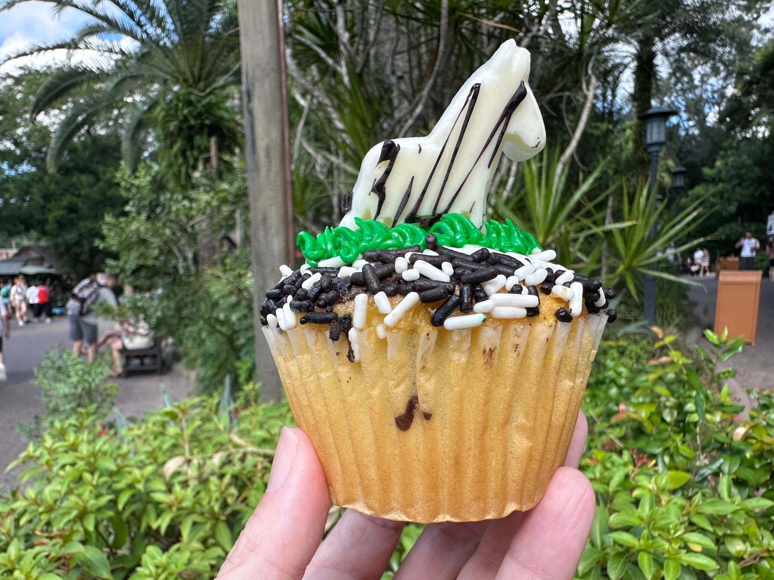A vanilla cupcake with white and black sprinkles, topped with green frosting and a white chocolate zebra figurine, held up in an outdoor setting reminiscent of Disney's Animal Kingdom, with trees and people in the background. Perfect for fans of Simba and Zebra Cupcakes alike.