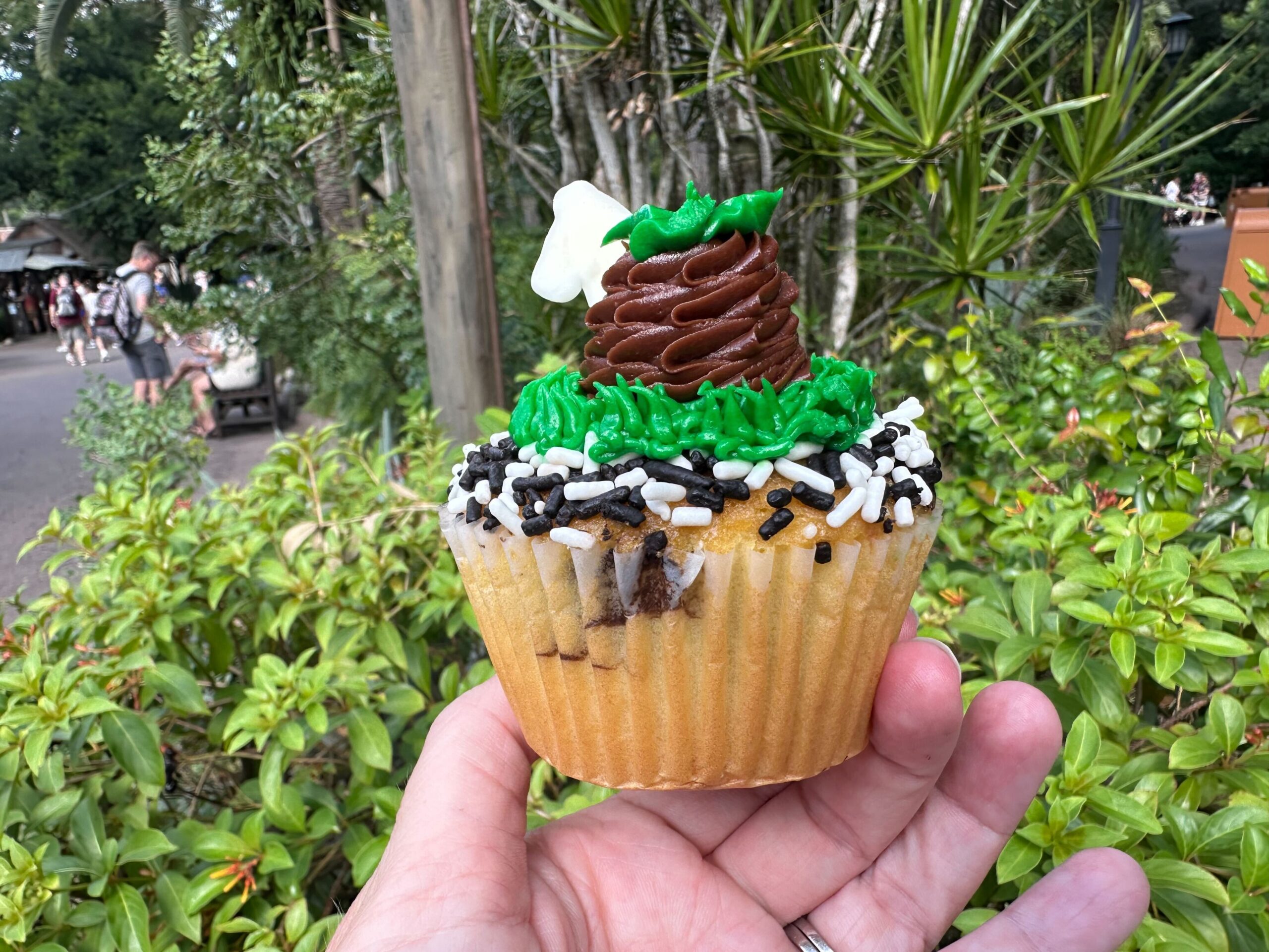 A person holds a cupcake with green icing grass, a chocolate mound, and a white bone decoration. Reminiscent of Zebra cupcakes found at Disney's Animal Kingdom, the treat is topped with black and white sprinkles, and it is set against a background of greenery.