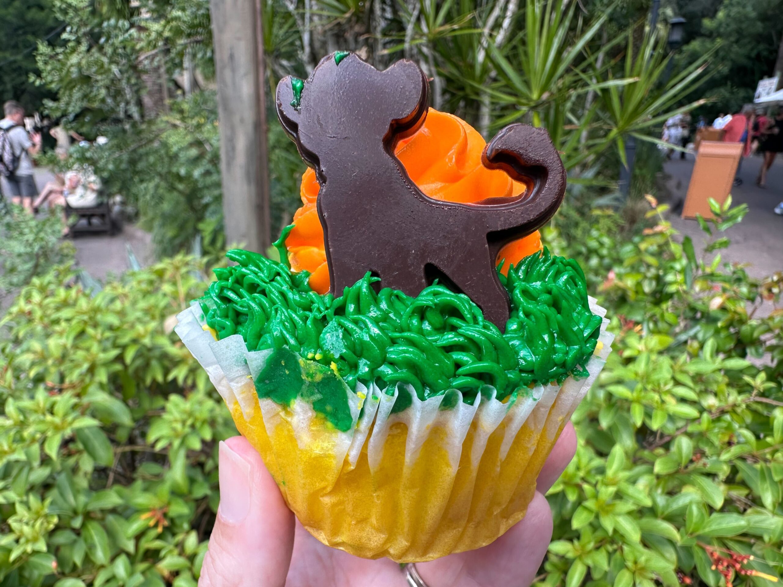 A hand holds a cupcake decorated with green and orange icing, topped with a chocolate silhouette of Simba. The background showcases outdoor foliage and people in the distance, evoking the ambiance of Disney's Animal Kingdom.