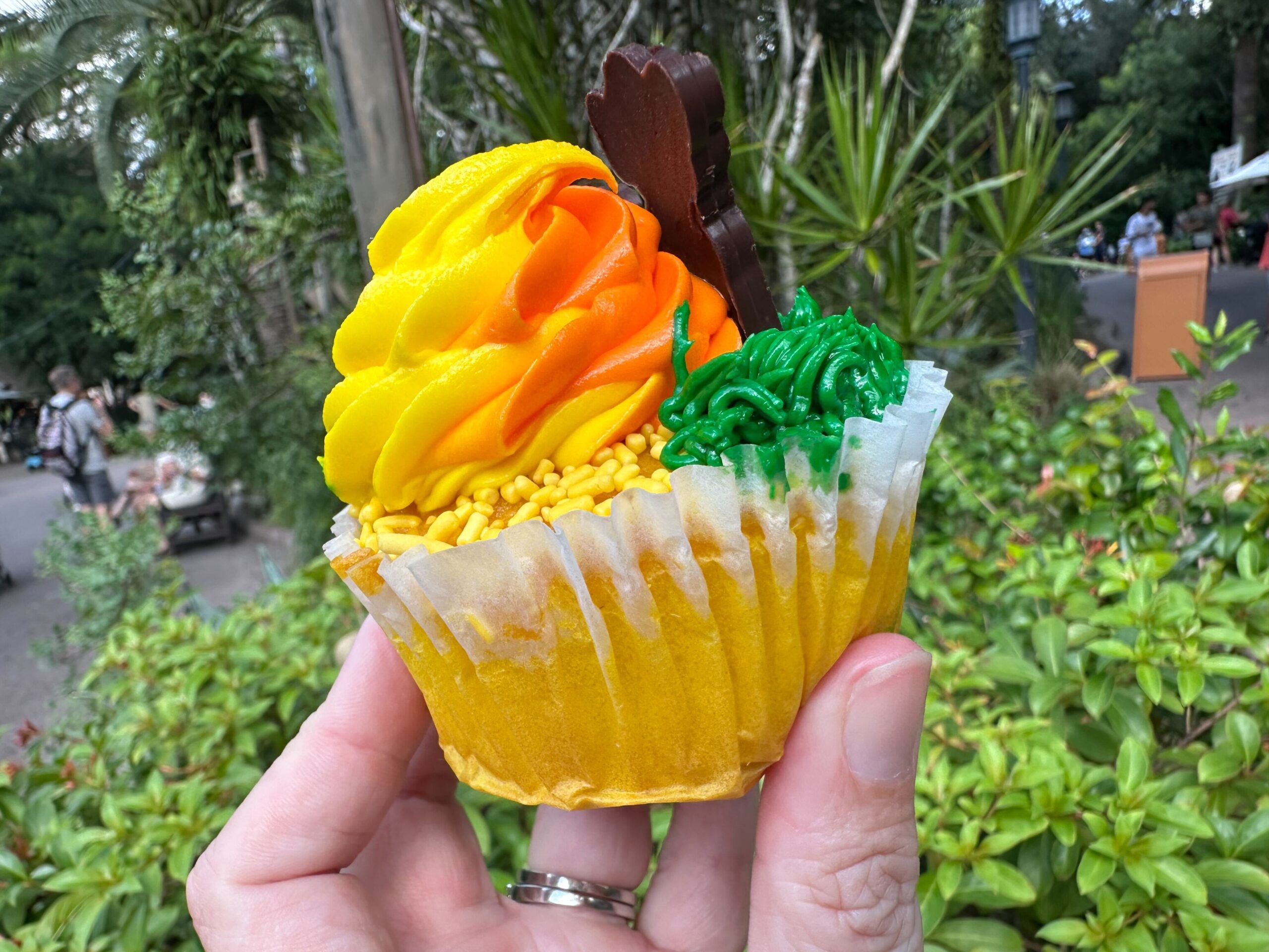 A hand holding a cupcake with yellow and orange frosting, green icing, and a small chocolate decoration on top. In the background, you can spot various plants and a pathway reminiscent of Disney's Animal Kingdom.