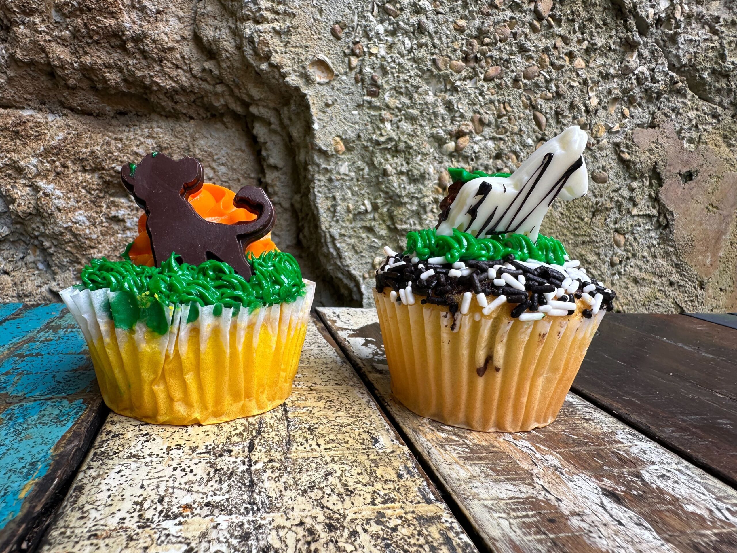 Two cupcakes on a rustic wooden surface: one with a chocolate lion-shaped topper and green icing, the other with a white zebra-shaped topper, green and black icing, and sprinkles.