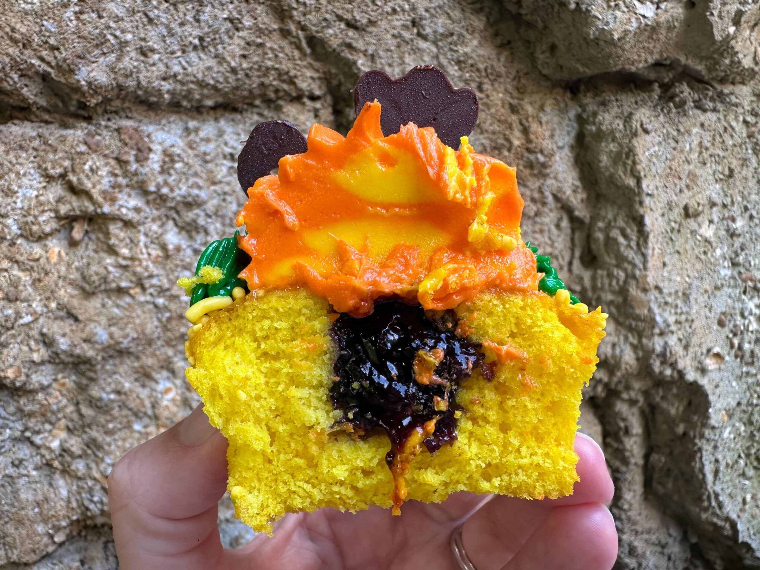 A person holds half of a yellow cupcake filled with dark jam and topped with orange frosting, reminiscent of one of the Zebra Cupcakes from Disney's Animal Kingdom, in front of a rough, beige stone background.
