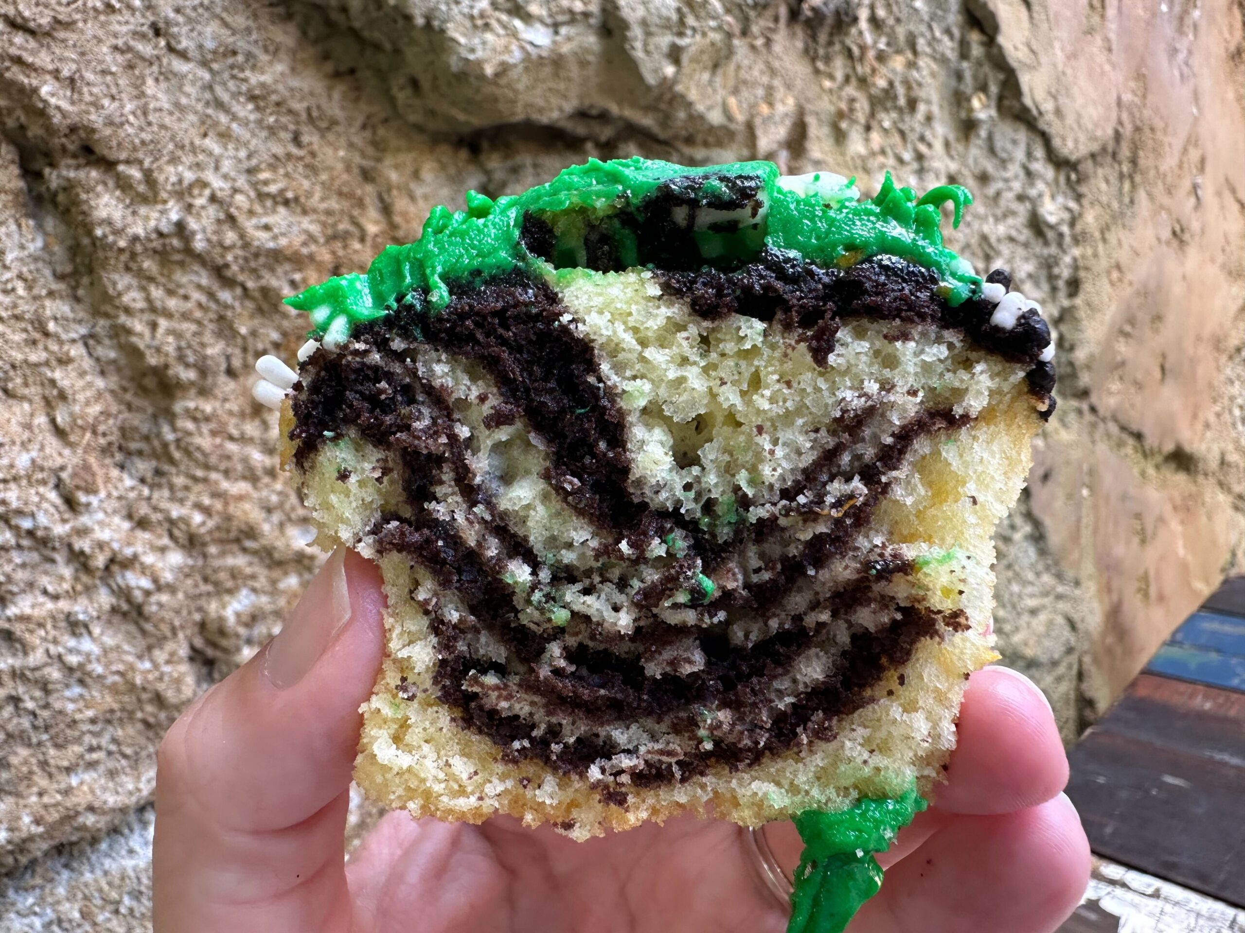 A hand holding a slice of marble cake with green frosting, revealing a pattern of chocolate and vanilla layers inside that resemble the iconic Zebra Cupcakes from Disney's Animal Kingdom. The background is a textured stone surface.