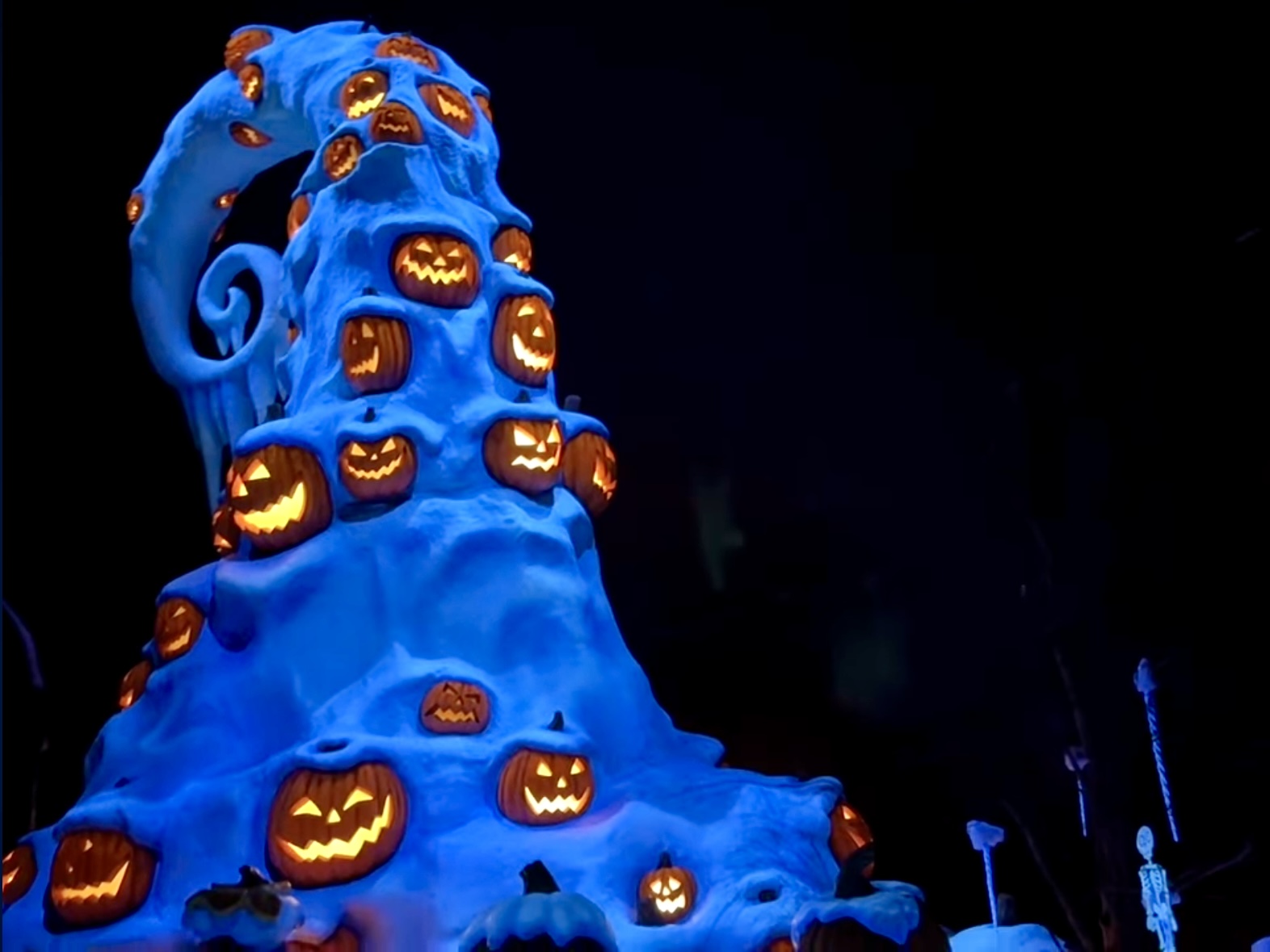 A towering, snow-covered spiral structure is decorated with orange jack-o'-lanterns, all illuminated with carved faces, set against a dark night sky, reminiscent of the enchanting atmosphere you might find at Tokyo Disneyland's Haunted Mansion Holiday Nightmare.