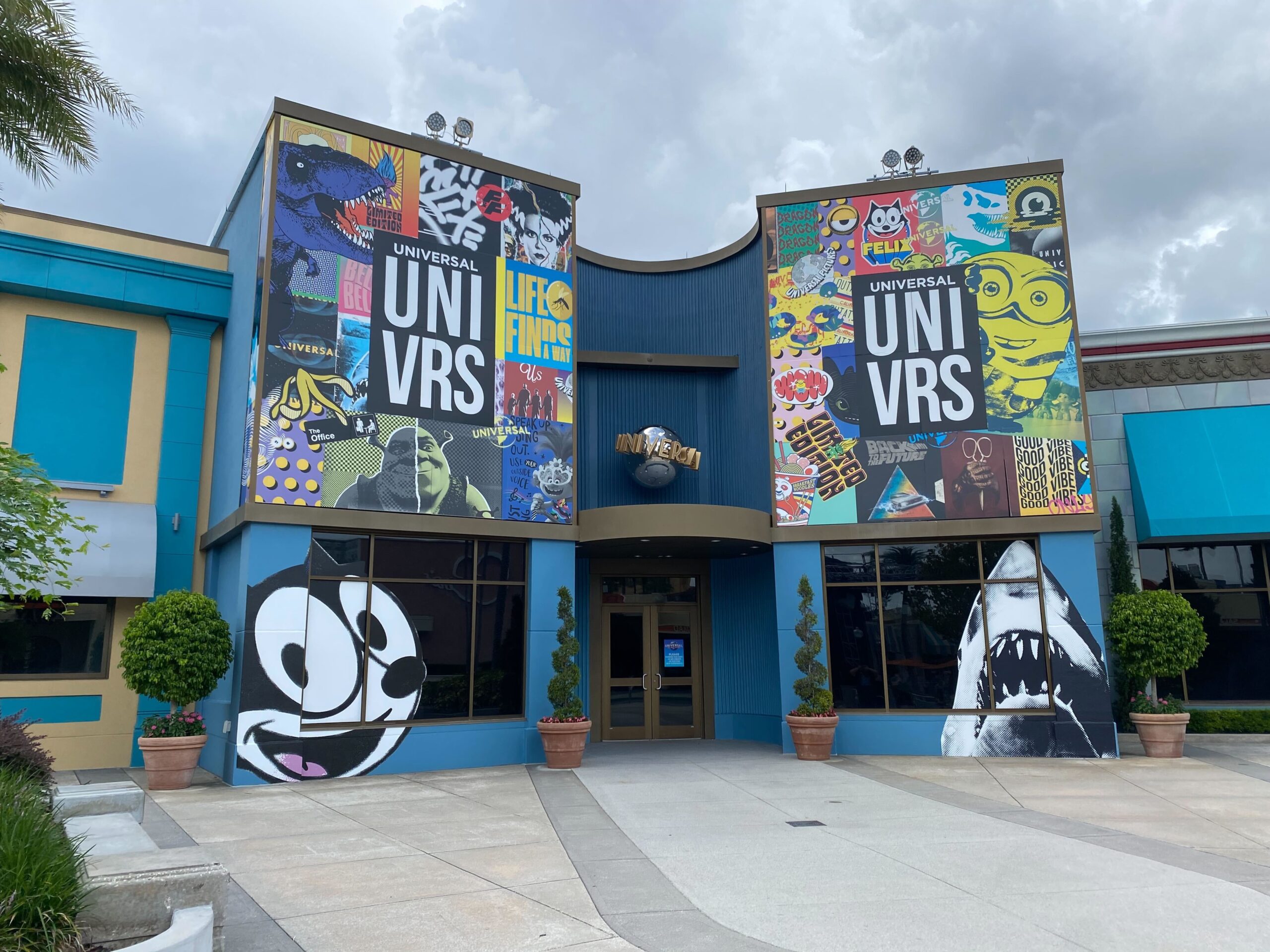 Entrance to the Universal Studios building decorated with colorful murals featuring various characters and themes. The building has two large banners with "Universal" text and posters on either side, including a vibrant Hello Kitty mural in the Uni vre Store section.