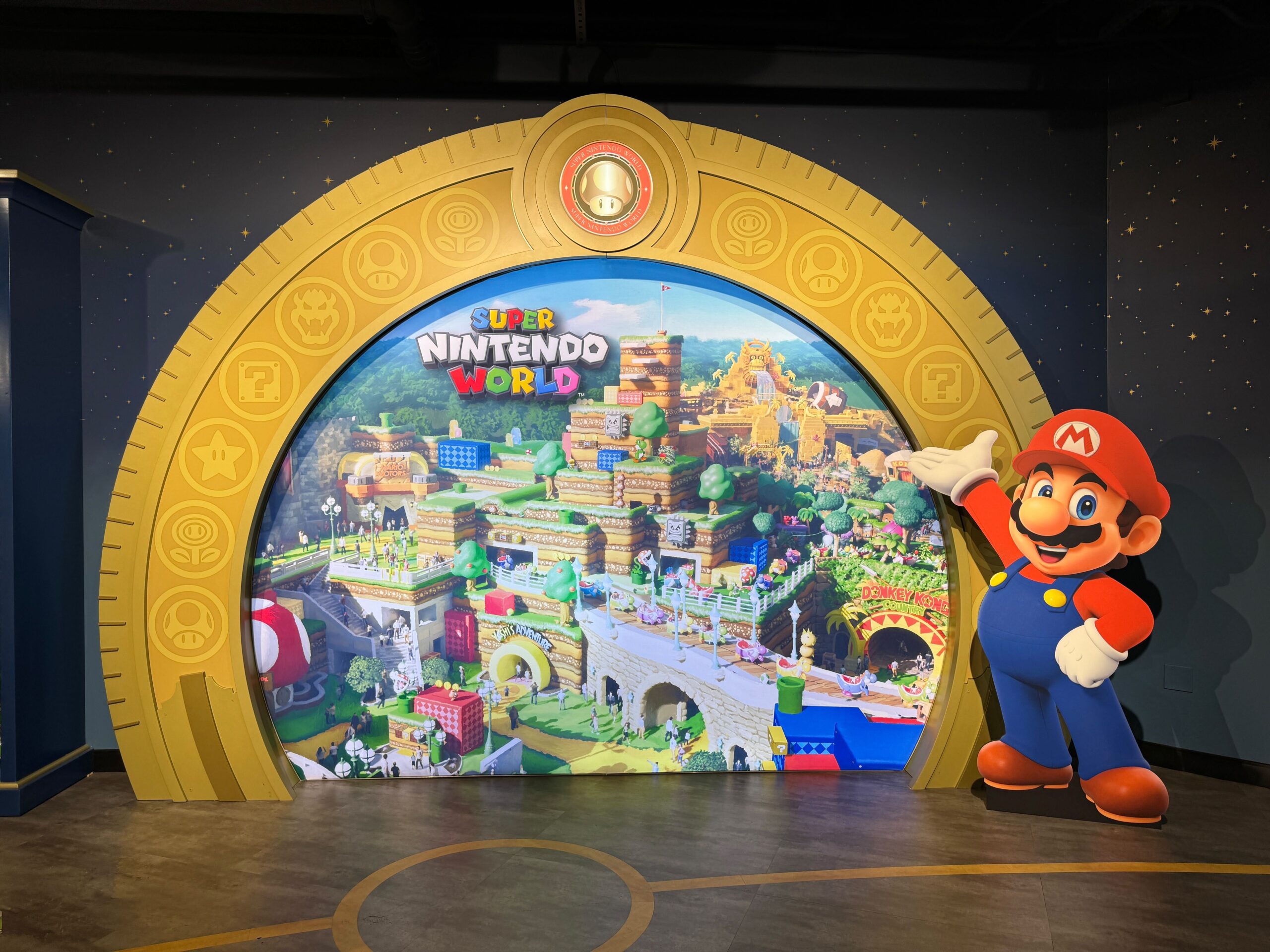 A colorful display of Super Nintendo World featuring a large image of the themed park. A life-size Mario figure stands on the right side, gesturing towards the background with an inviting pose.