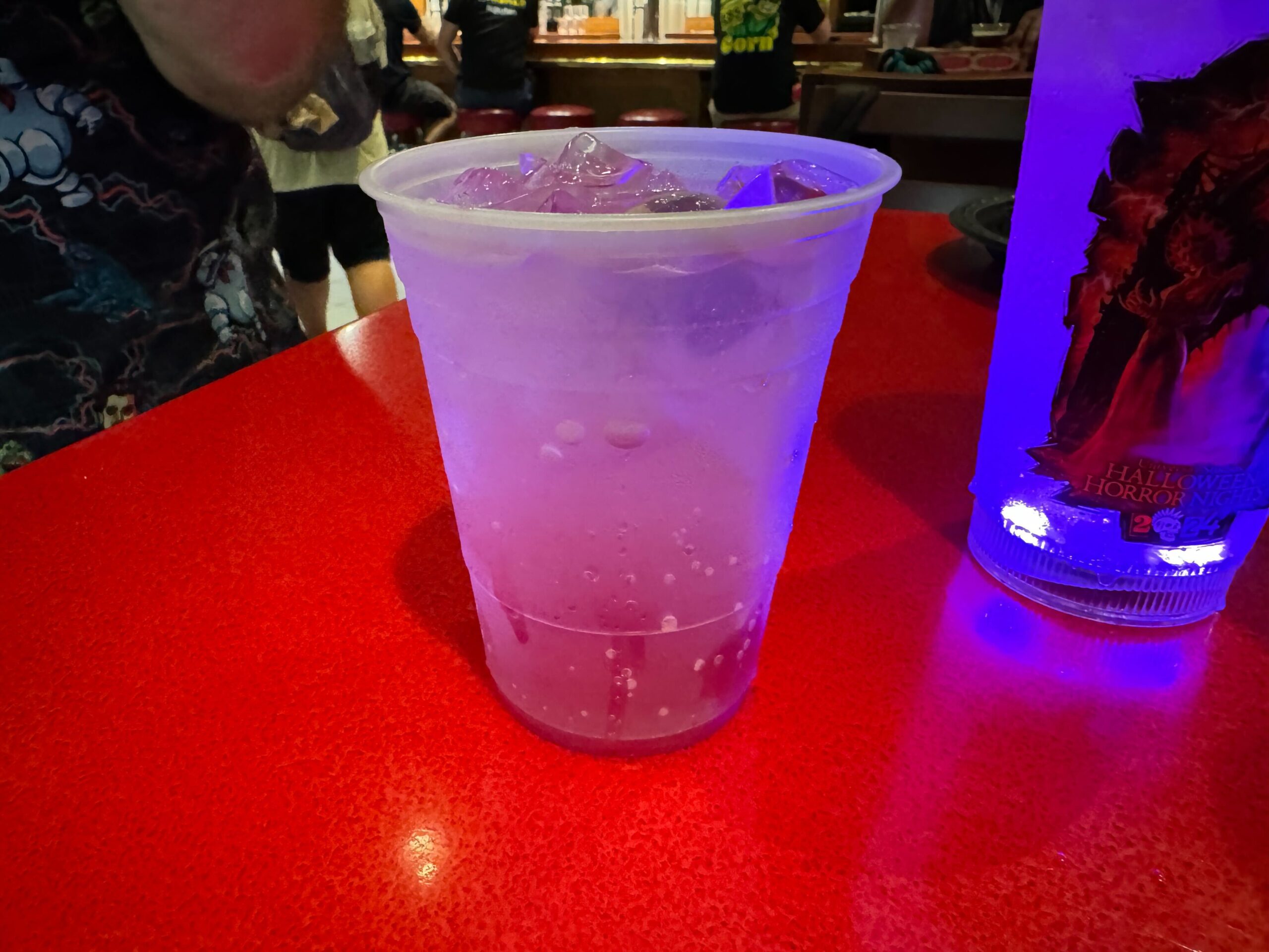 A clear plastic cup filled with a purple liquid and ice sits on a glossy red table, perfect for those wild nights at Halloween Horror Nights 33. An illuminated drink container with graphics is partially visible on the right side, inviting guests to eat and drink amidst the spooky festivities.
