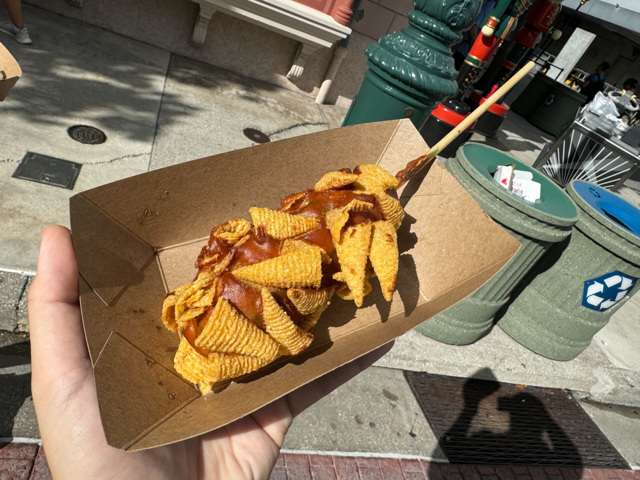 A person holds a cardboard tray containing a skewered snack, covered in crispy corn chips, with a street scene and recycling bin in the background. Perfect for those reviews on local eats or while enjoying events like Halloween Horror Nights.