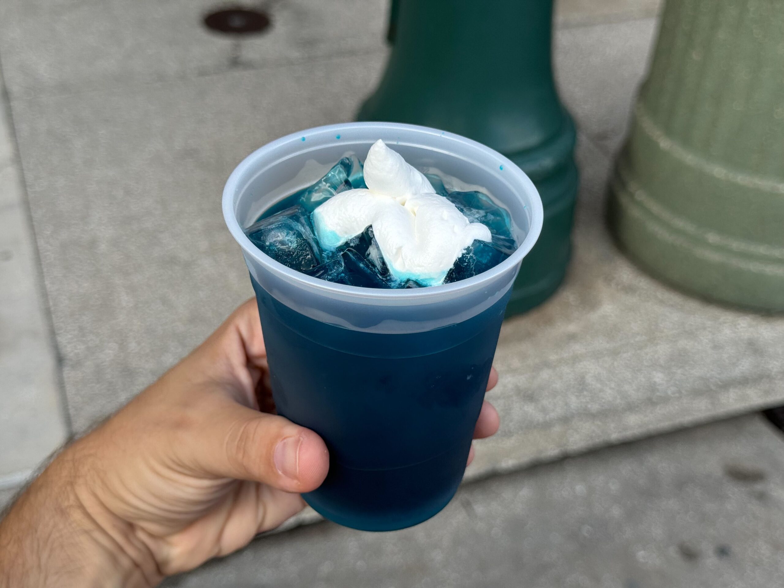 A hand holds a plastic cup filled with blue liquid, ice cubes, and a white flower-shaped garnish—a perfect treat for Everything You Can Eat & Drink at Halloween Horror Nights.