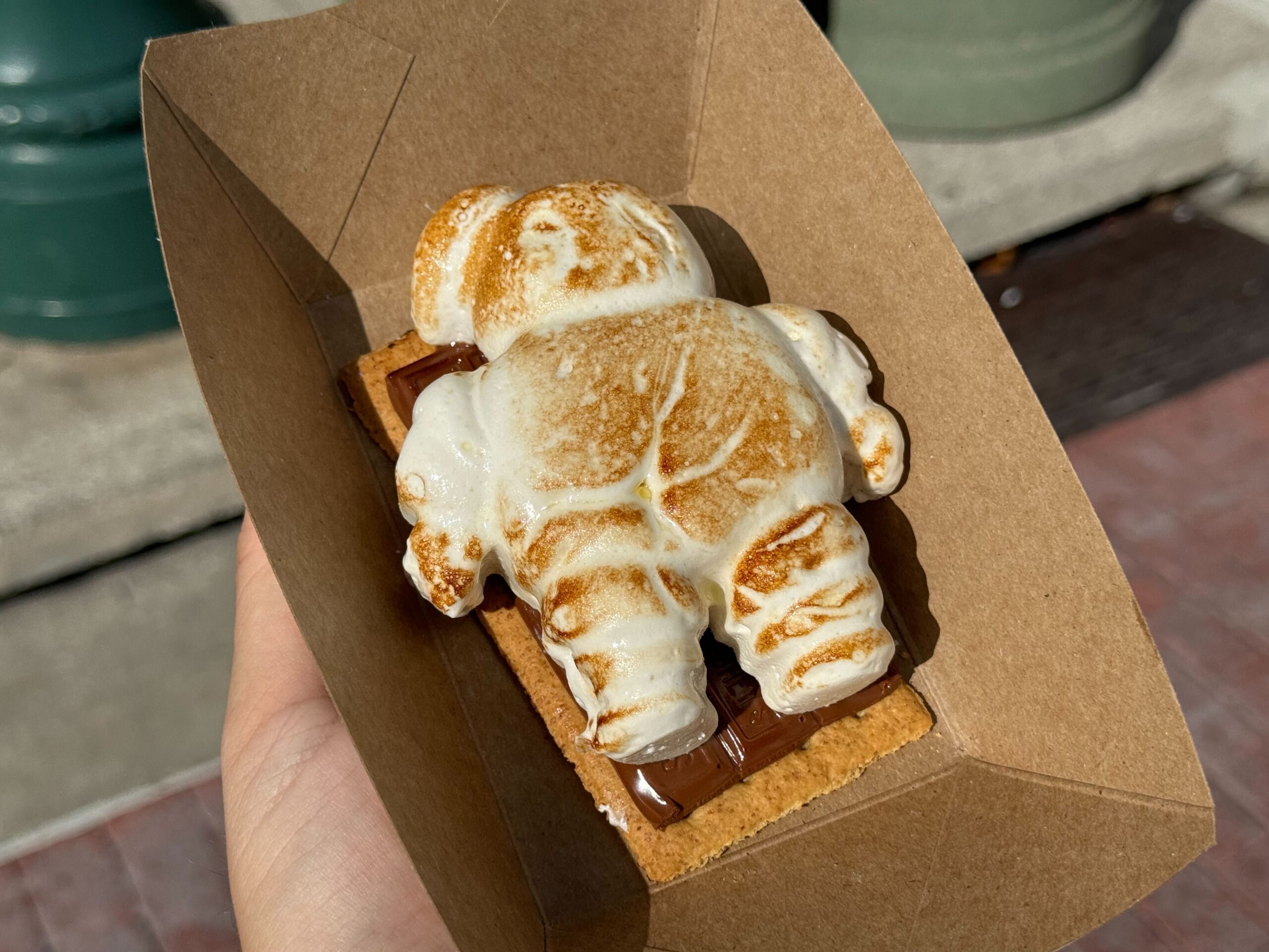 A roasted marshmallow figure resembling a character lies on a chocolate and graham cracker base, creating a unique s'more, held in a brown paper container. This delightful treat is perfect for those enjoying the spooky ambiance of Halloween Horror Nights.