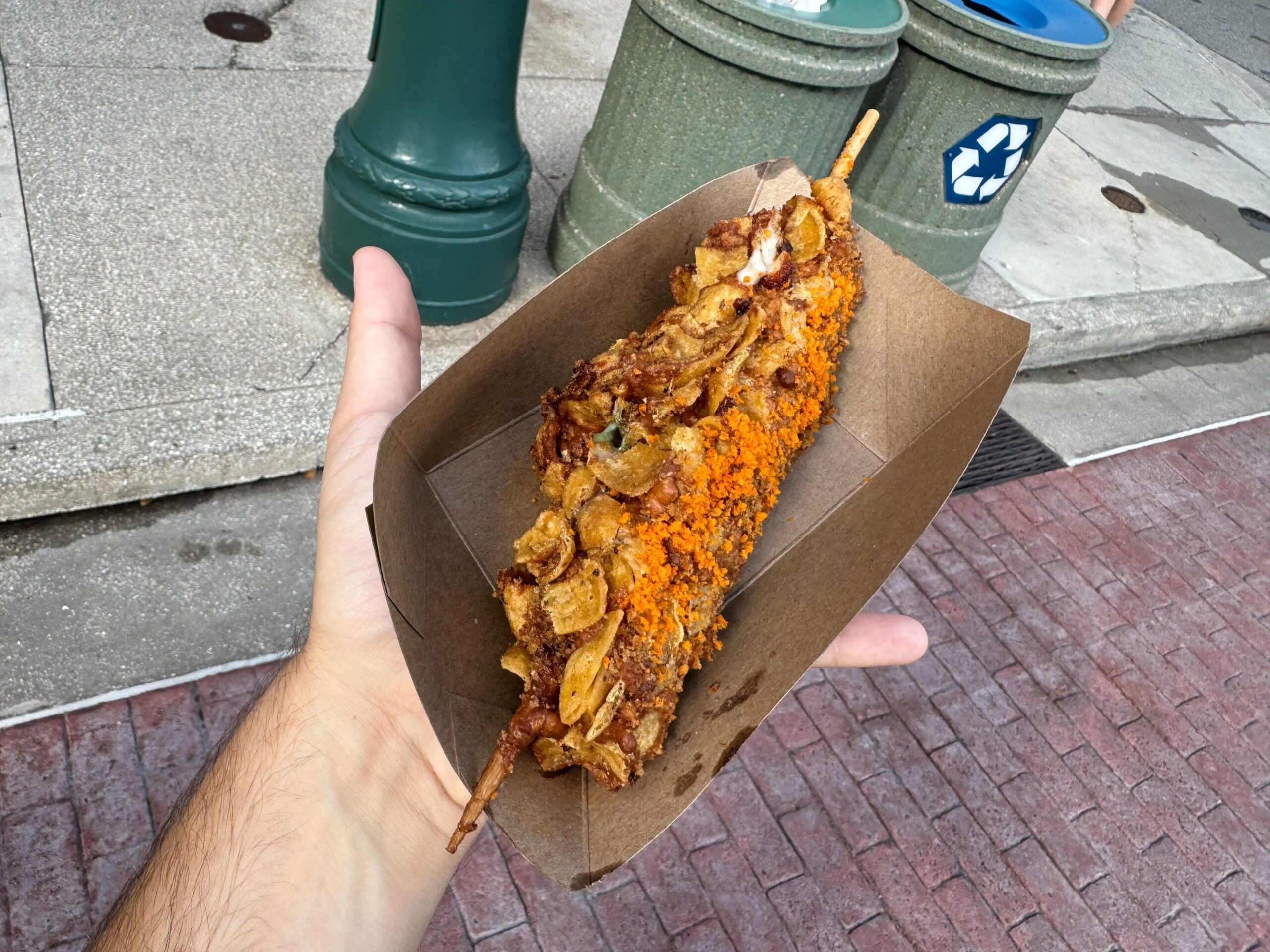 A person holds a fried, skewered food item topped with various seasonings and bits in a cardboard tray, with a street, trash bins, and a green post visible in the background—perfect for an Eat & Drink review during Halloween Horror Nights.