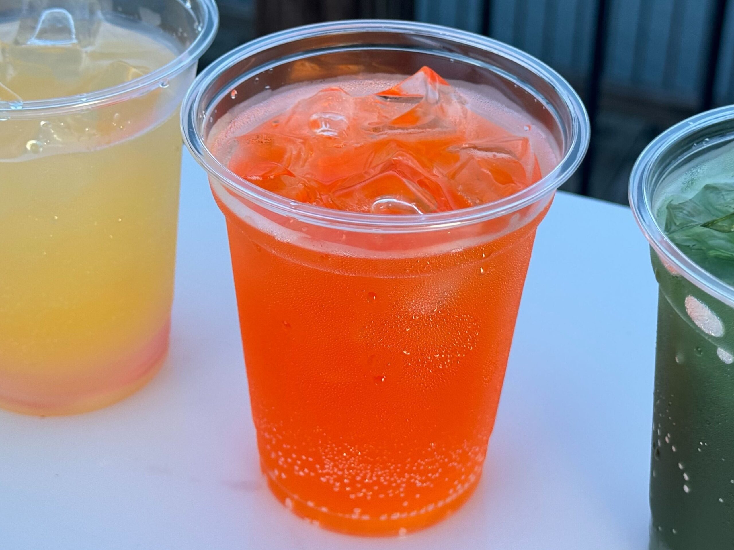 Three plastic cups of beverages on a white surface at Halloween Horror Nights; one with a yellow drink, one with an orange drink, and one with a green drink. Eat & Drink and enjoy the spooky ambiance!