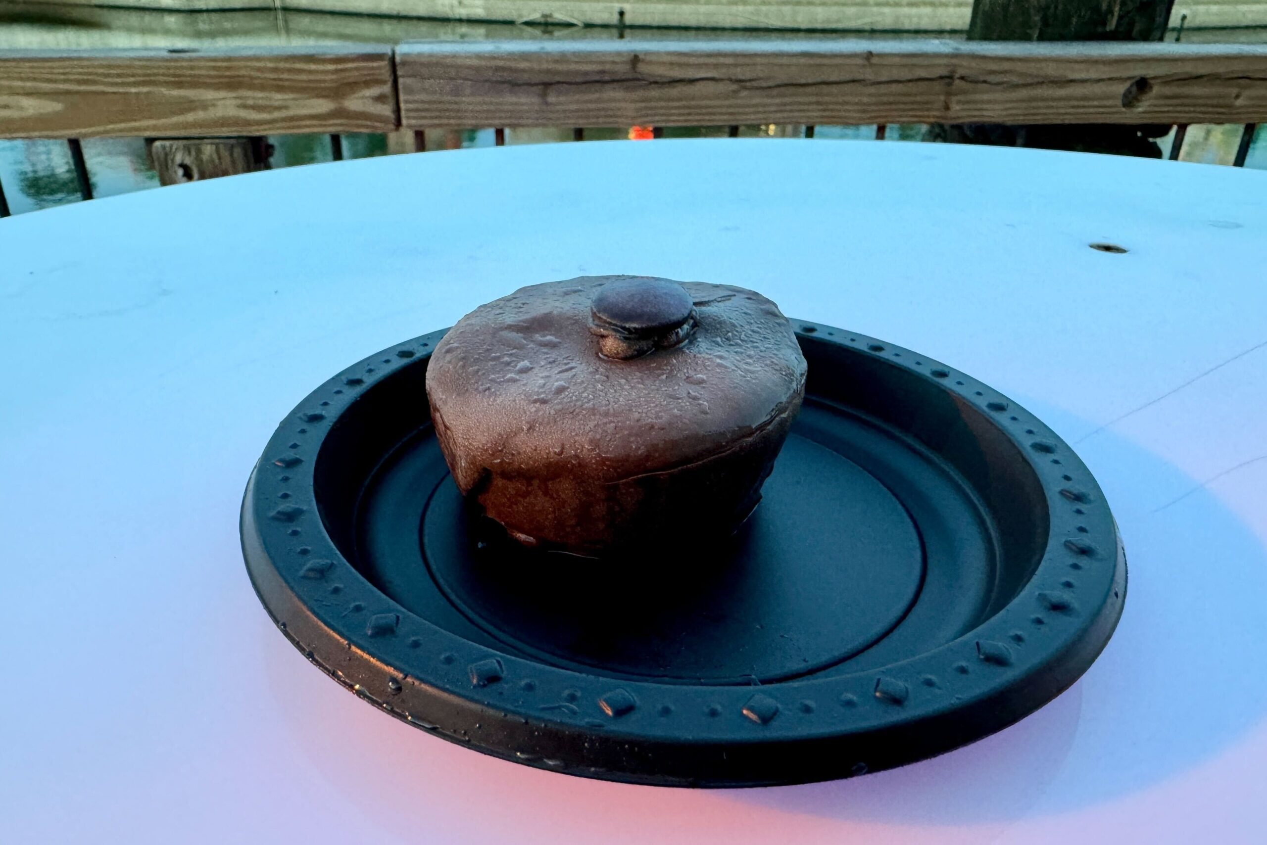 A small, round, chocolate-covered dessert with a chocolate disk on top, placed on a decorative black plate on an outdoor table. A fence and water are visible in the background, making it an ideal treat to enjoy while reading reviews about Halloween Horror Nights.